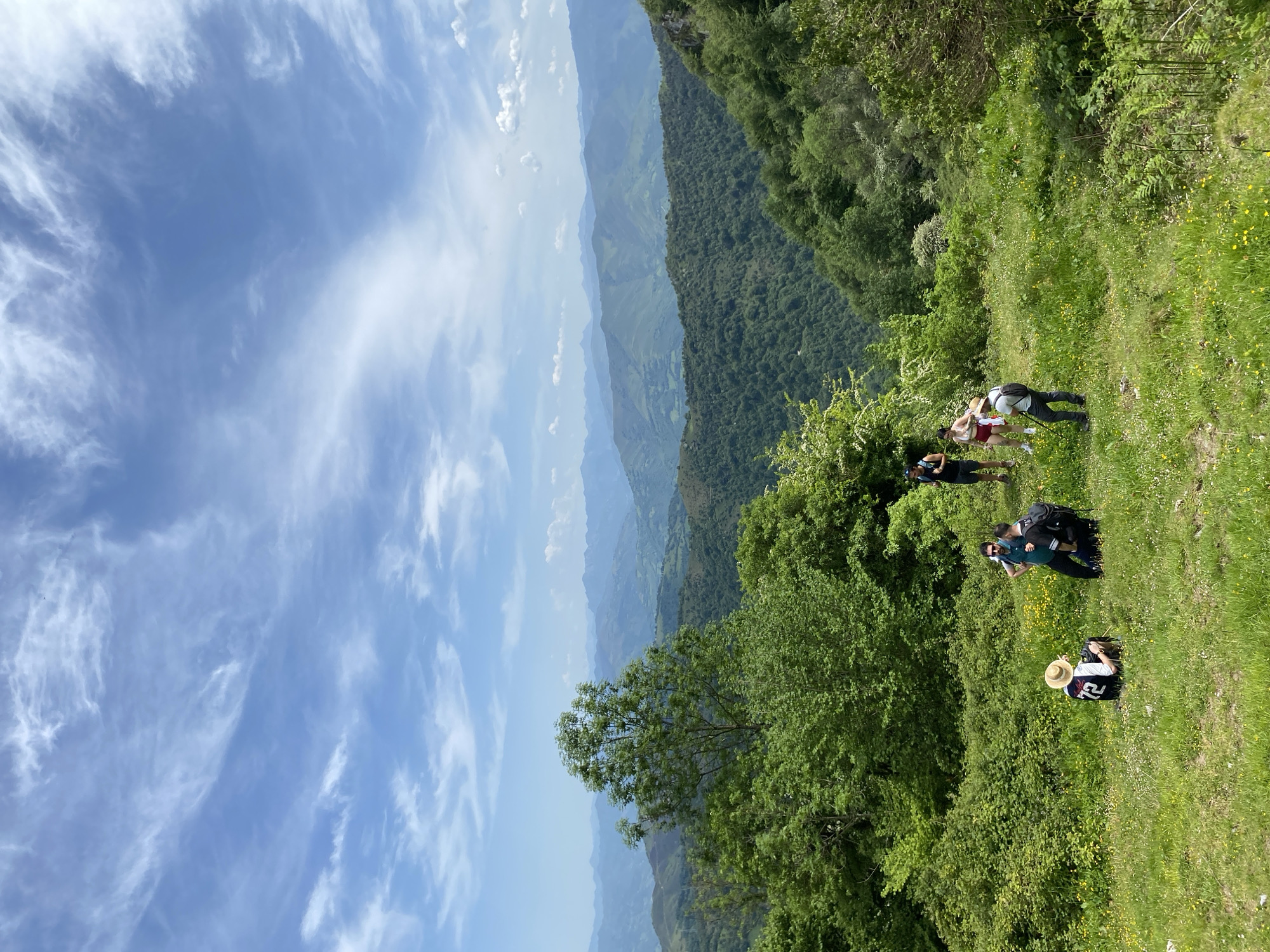 The image captures a group of people on a grassy hillside in what appears to be a mountainous region, under a blue sky with wispy clouds. The location may be Riosa, Spain, based on the prompt. In the foreground, a person wearing a straw hat and a jersey with the number "72" sits on the grass. A small group of people stand behind and slightly above this person. Behind the group are more people who are standing. The hillside is covered in grass and wildflowers. In the background, a series of mountain ranges fade into the distance. The scene is dominated by green hues from the vegetation, contrasted by the blue sky and the distant mountains.