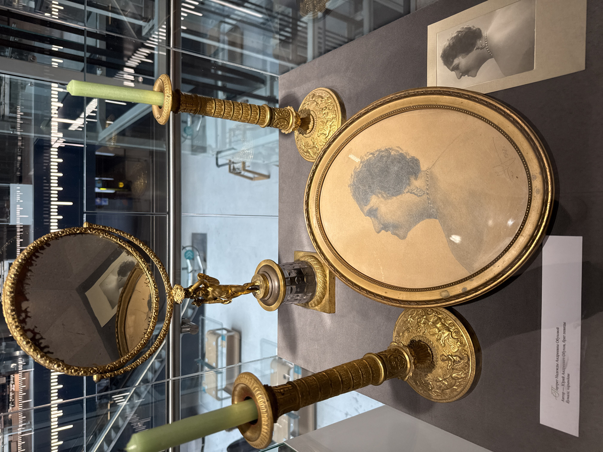 The image depicts a still life arrangement featuring an oval framed portrait of a woman, a gold-plated candelabra with a green candle, a gold-plated hand mirror with a winged cherub figurine at its base, another gold-plated candelabra with a green candle, and a small black and white photograph of a woman. The items are displayed on a dark gray surface, likely a museum display case. The background shows a modern interior with glass partitions and what appear to be escalators, suggesting a public exhibition space. A descriptive plaque is visible in the lower left corner with Russian text. The text on the plaque reads "Портрет Надежды Андреевны Обуховой, Автор – Юрий Андреевич Обухов, брат певицы. Бумага, карандаш" which translates to "Portrait of Nadezhda Andreevna Obukhova, Author – Yuri Andreevich Obukhov, singer's brother. Paper, pencil." The overall impression is one of historical display and artistic presentation. The scene does not provide specific location details within Moscow.