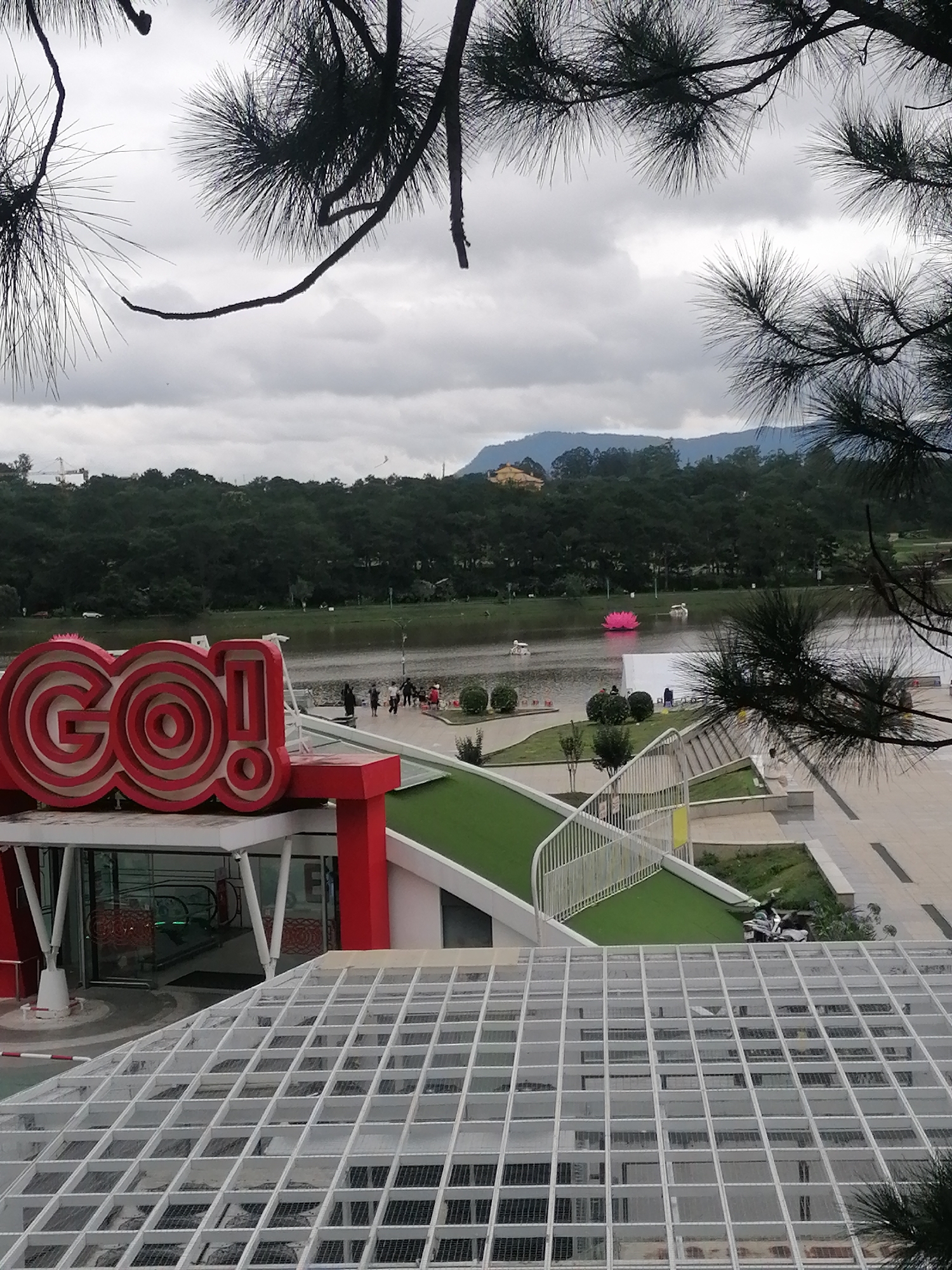 The photograph shows a view of Xuan Huong Lake in Da Lat, Vietnam. In the foreground is the "GO!" hypermarket, identifiable by its large red sign. The lake features several swan-shaped paddle boats and a large, artificial pink lotus flower. A group of people are gathered on the bank near the lake. The background includes a line of trees and hills under an overcast sky. Pine tree branches frame the top of the image. A pedestrian overpass is seen along the lakeshore.