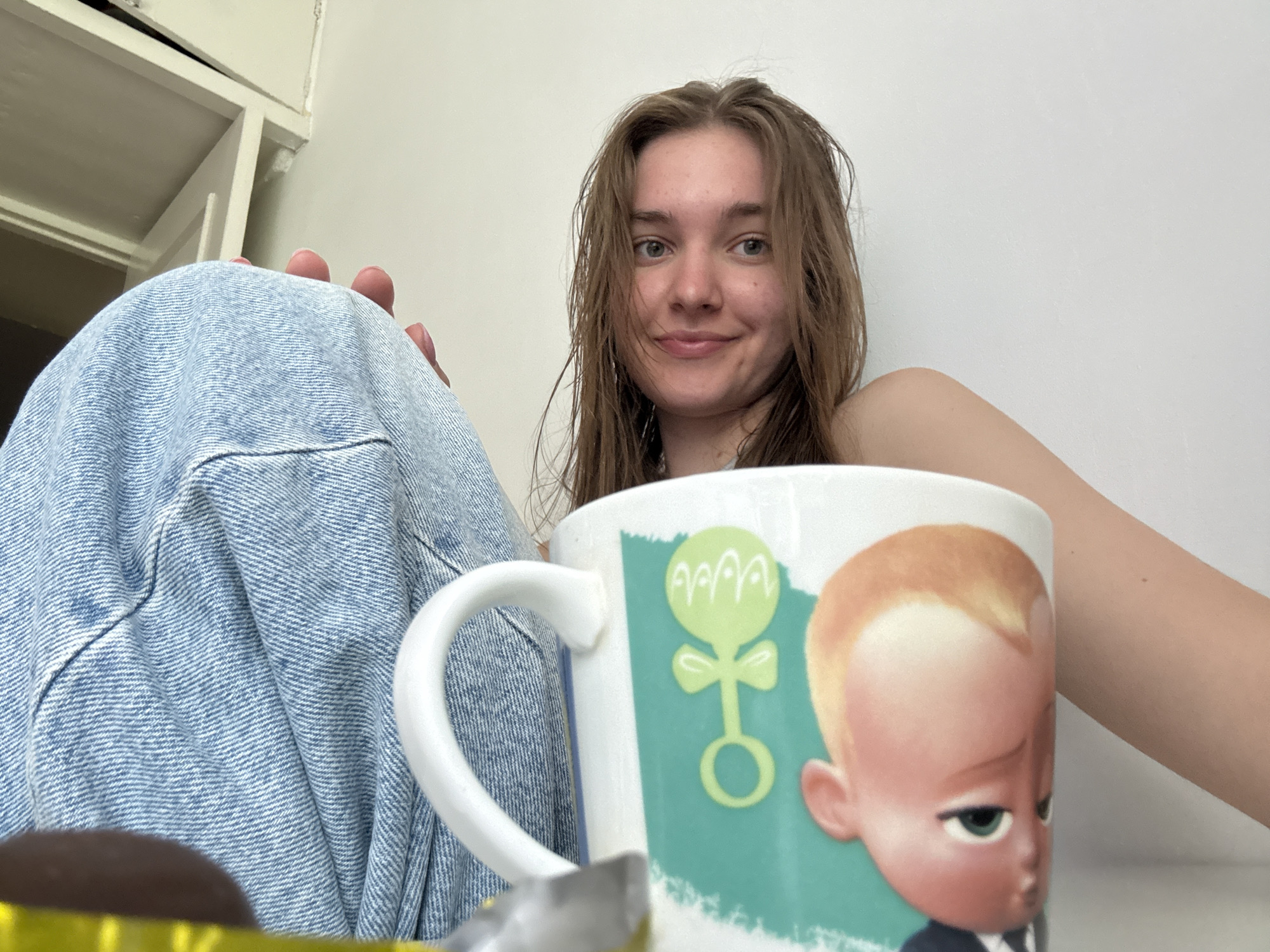 A young woman is sitting indoors, likely in a residential setting. She has wet, light brown hair and is smiling slightly at the camera. She is wearing light blue denim jeans. A white mug with a cartoon image of the "Boss Baby" is prominently displayed in the foreground. The mug features a light green background with a rattle design. Part of a spoon is visible within the mug, along with a piece of something wrapped in gold foil. A white wall is behind her. The location is unknown.