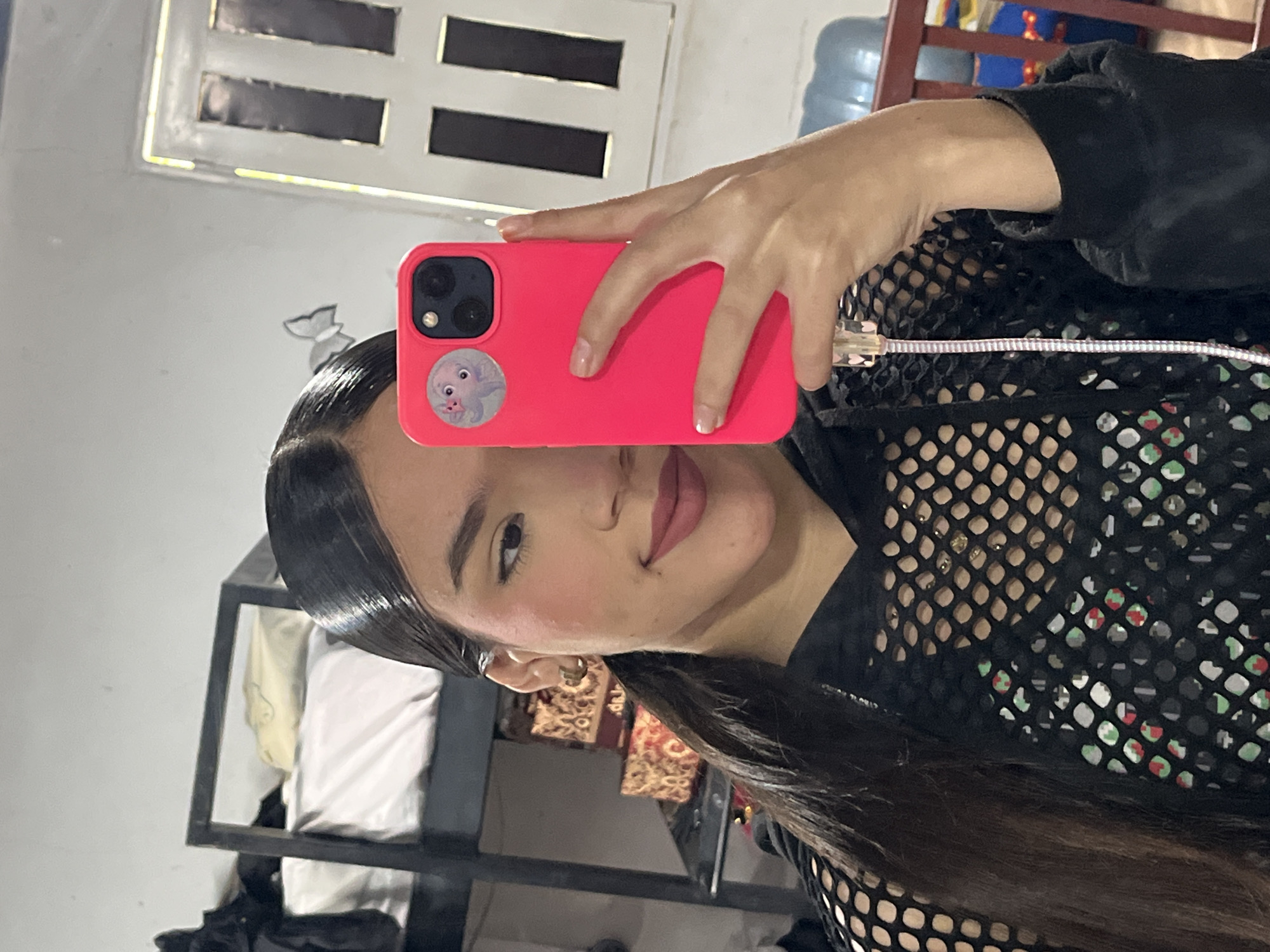 In an indoor setting, possibly a bedroom or living area, a young woman with dark hair pulled back in a ponytail takes a mirror selfie. She is wearing a black mesh top over a printed shirt. Her right hand holds a bright pink smartphone with a cartoon sticker on the case. The background shows a bunk bed, some patterned boxes on a shelf, and a window with vertical panes. A charging cable is connected to the phone. The environment appears somewhat cluttered, suggesting a personal living space. The image is captured in Juan Griego, Venezuela.