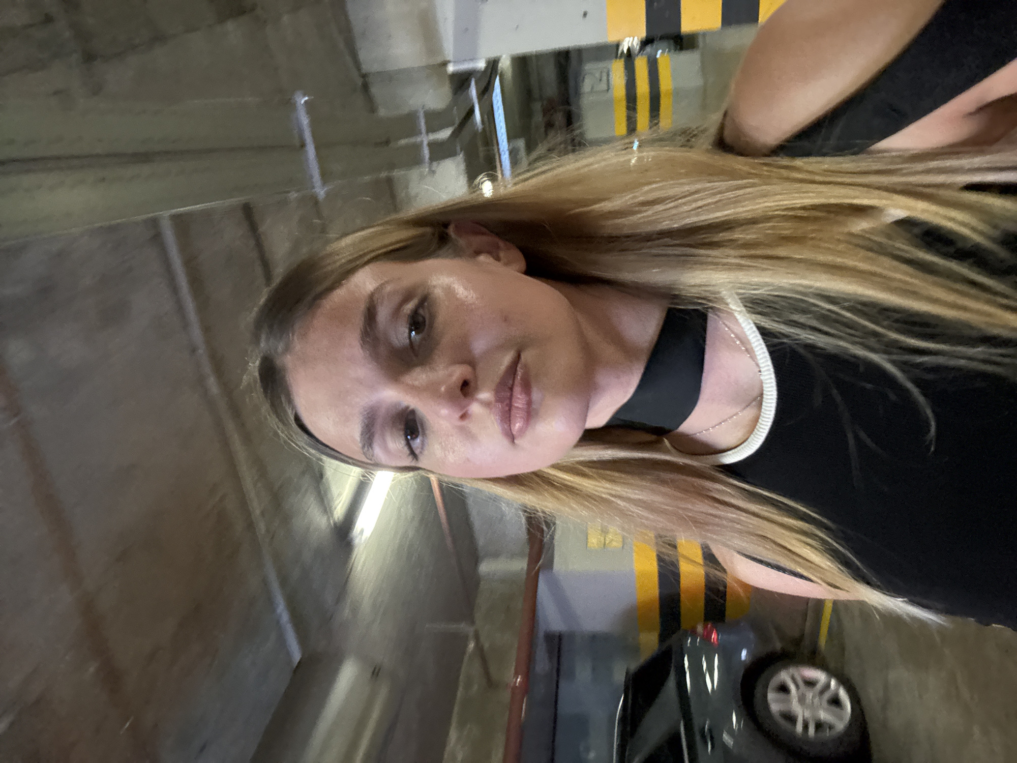 In a dimly lit underground parking garage, a young woman with long blonde hair stands facing the camera, taking a selfie. She is wearing a black tank top with a white trim and a black choker necklace. Her expression is neutral, and her gaze is directed towards the camera. The background shows the concrete ceiling and walls of the garage, along with a parked car and yellow and black striped barriers. The lighting suggests artificial illumination from overhead fluorescent lights.