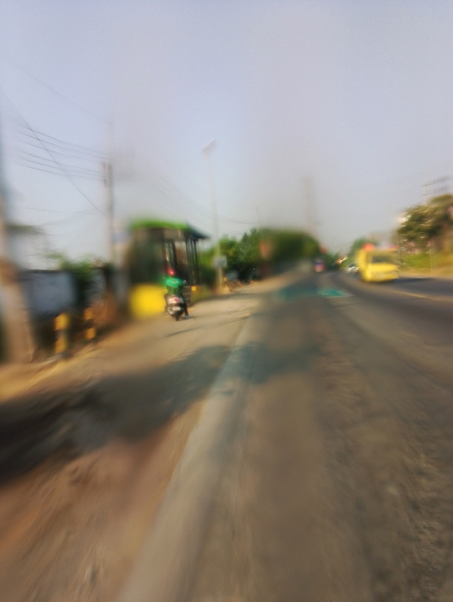 The image, captured from a moving vehicle, depicts a street scene with a shallow depth of field and motion blur. On the left side of the road, a bus stop shelter with a green roof is visible, with a scooter parked in front of it. The scooter has a rider wearing a green helmet and jacket. The road itself is asphalt, with a yellow bus visible further down on the right. Trees and buildings line the sides of the road, and utility poles with wires stretch across the sky. The overall impression is one of a bustling urban environment, viewed from a dynamic perspective.