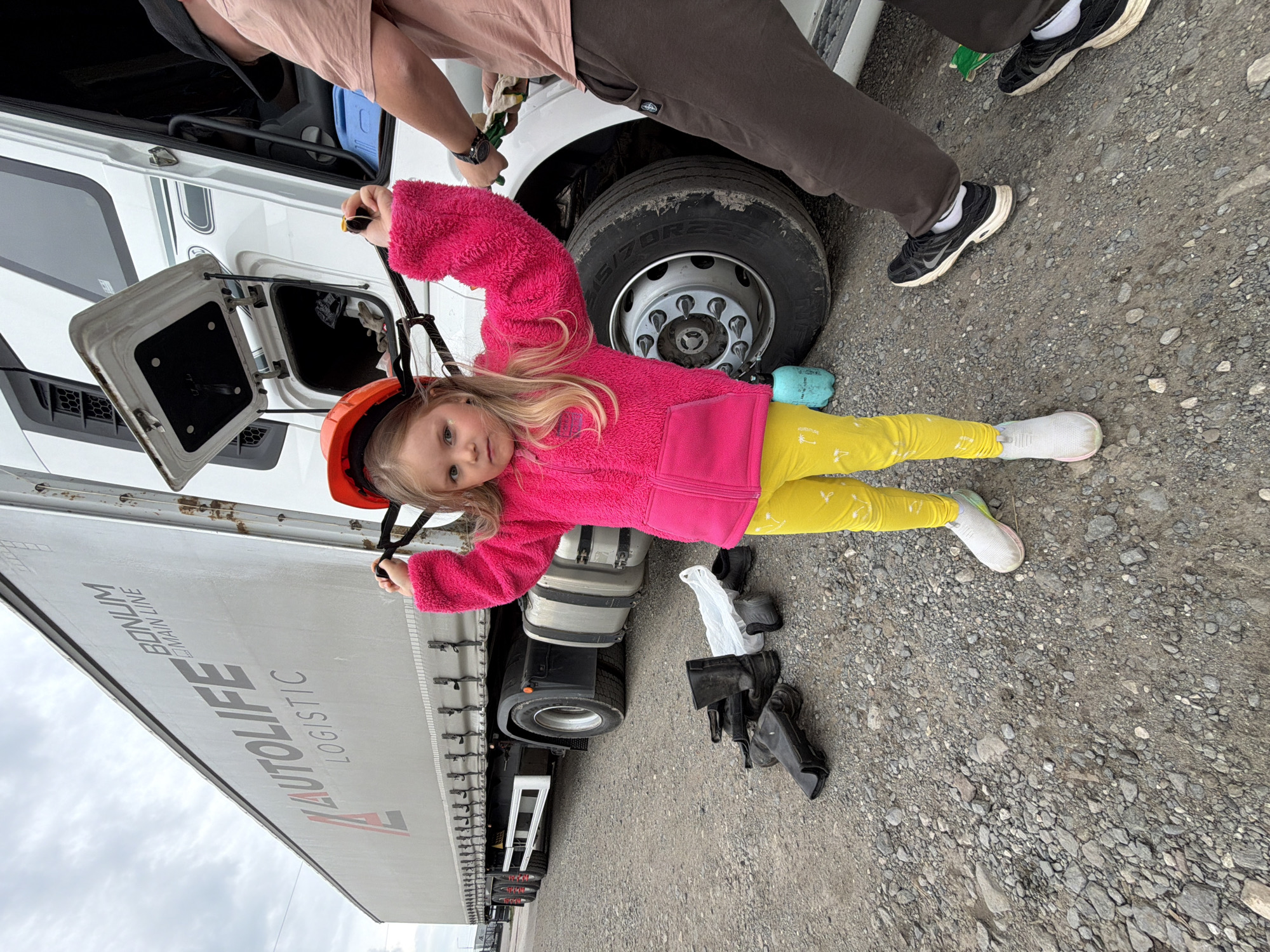 An outdoor scene in Kamyennoye, Russia, shows a young girl lying on a gravel surface next to a large white semi-truck and its trailer. The girl has long blonde hair and is wearing a bright pink fuzzy jacket, yellow leggings with faint patterns, white socks, and white sneakers. She has an orange helmet strapped to her head and holds onto a strap with both hands, looking directly at the camera with a neutral expression.
Behind her, the lower half of an adult's body is visible, wearing dark pants and black sneakers, standing next to a large truck tire (which displays "70R22.5" on its sidewall). Another person's arms are visible near the truck's cabin, seemingly engaged in an activity like refueling or maintenance, with a compartment open and a blue container visible inside. Various black, possibly plastic, vehicle parts or tools, and a white plastic bag are scattered on the gravel near the truck's rear.
The truck's trailer displays "AUTOLIFE LOGISTIC" prominently, with "BONUM MAIN LINE" written above it. The sky above is overcast, suggesting a cloudy daytime. The setting appears to be a truck stop or a roadside area, indicating a pause during transport.
