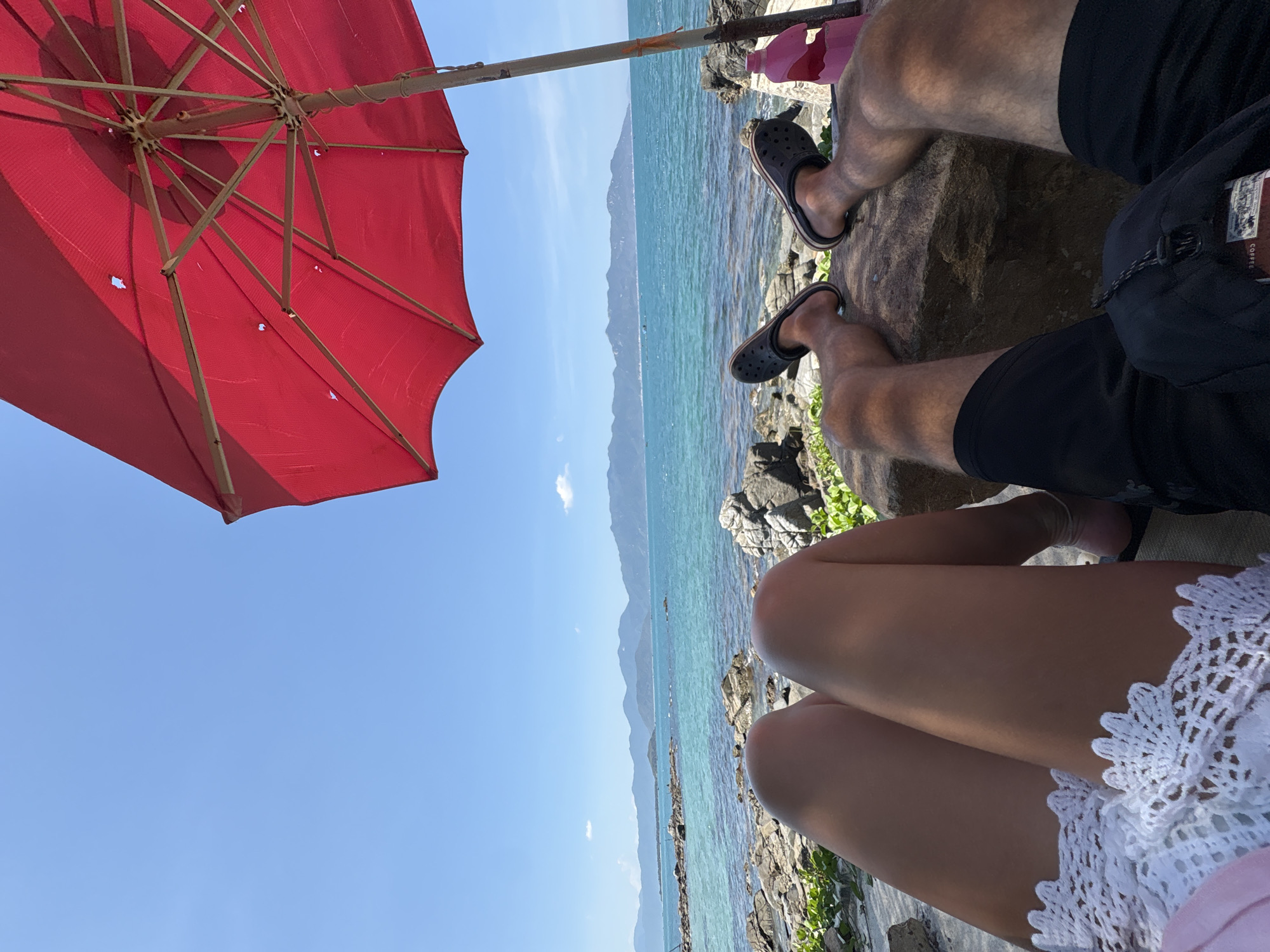 The image captures a sunny day at what appears to be a rocky coastline in Vĩnh Lương, Vietnam. The scene is framed by a red umbrella partially visible in the upper left corner, providing shade. The aqua sea and distant mountains form the backdrop. Two individuals are present. One individual is wearing black shorts and black clogs, sitting on a rock. Another individual, wearing a white crochet garment, is positioned in the lower part of the frame. The focus is on their legs and the immediate rocky environment. The scene suggests relaxation and a beachside setting.