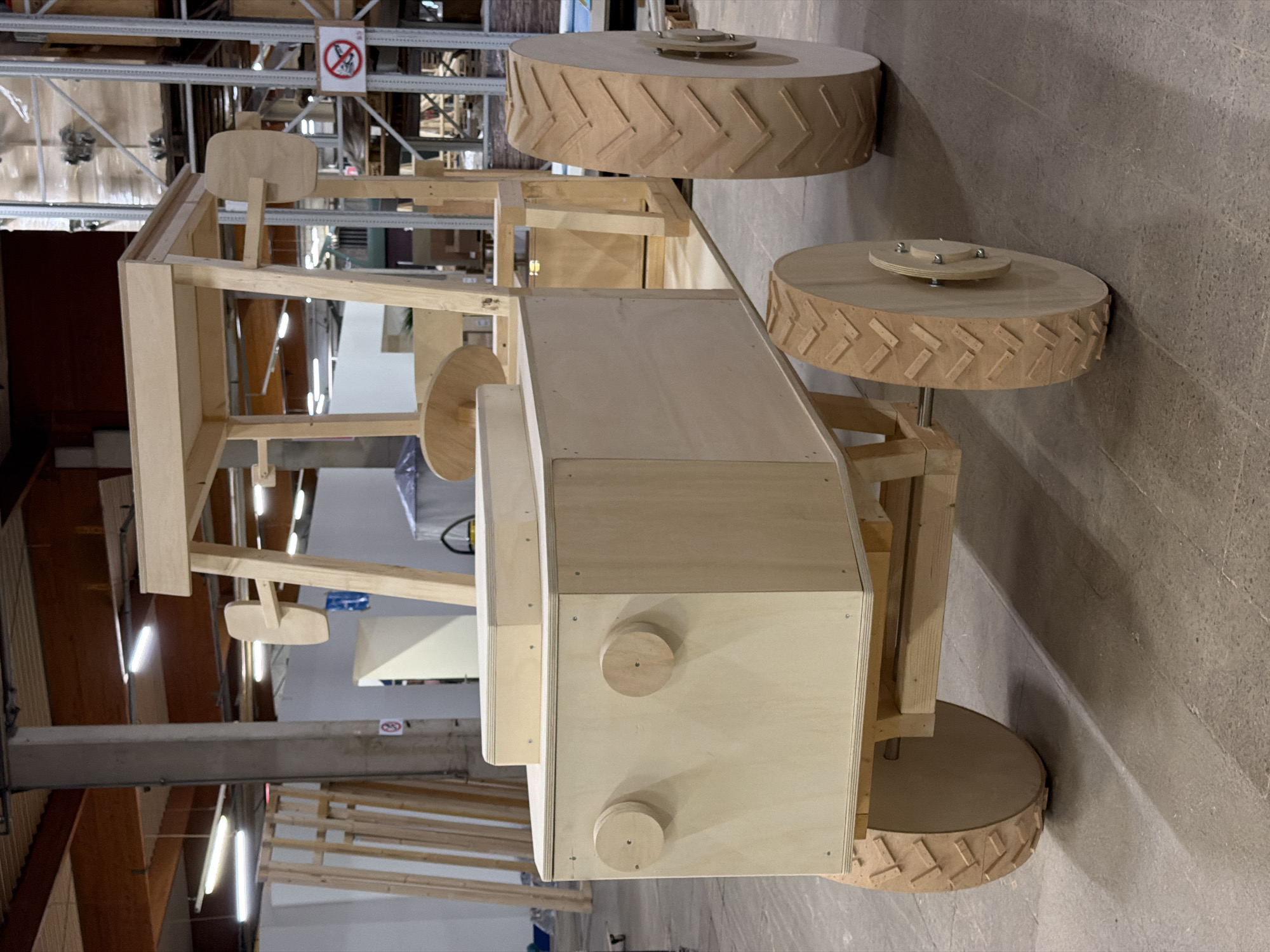 This is a full-shot, eye-level, medium-angle, indoor photograph of a wooden toy tractor. The tractor is unfinished, made of light-colored plywood and solid wood pieces. It features a large box-like body with two circular wooden knobs resembling headlights, and a smaller rectangular section on top. The tractor has a canopy structure supported by wooden posts, with two wooden steering wheels visible, one inside the main body and one attached to the canopy structure. It has four wooden wheels: two smaller ones at the front and two larger ones at the rear, all with carved tire treads. The tractor is positioned on a concrete floor. In the background, there are industrial shelving units stacked with various materials, wooden frames, fluorescent lights on the ceiling, and some signage. The overall environment appears to be a workshop or warehouse. The image was taken in Leudelange, Luxembourg.