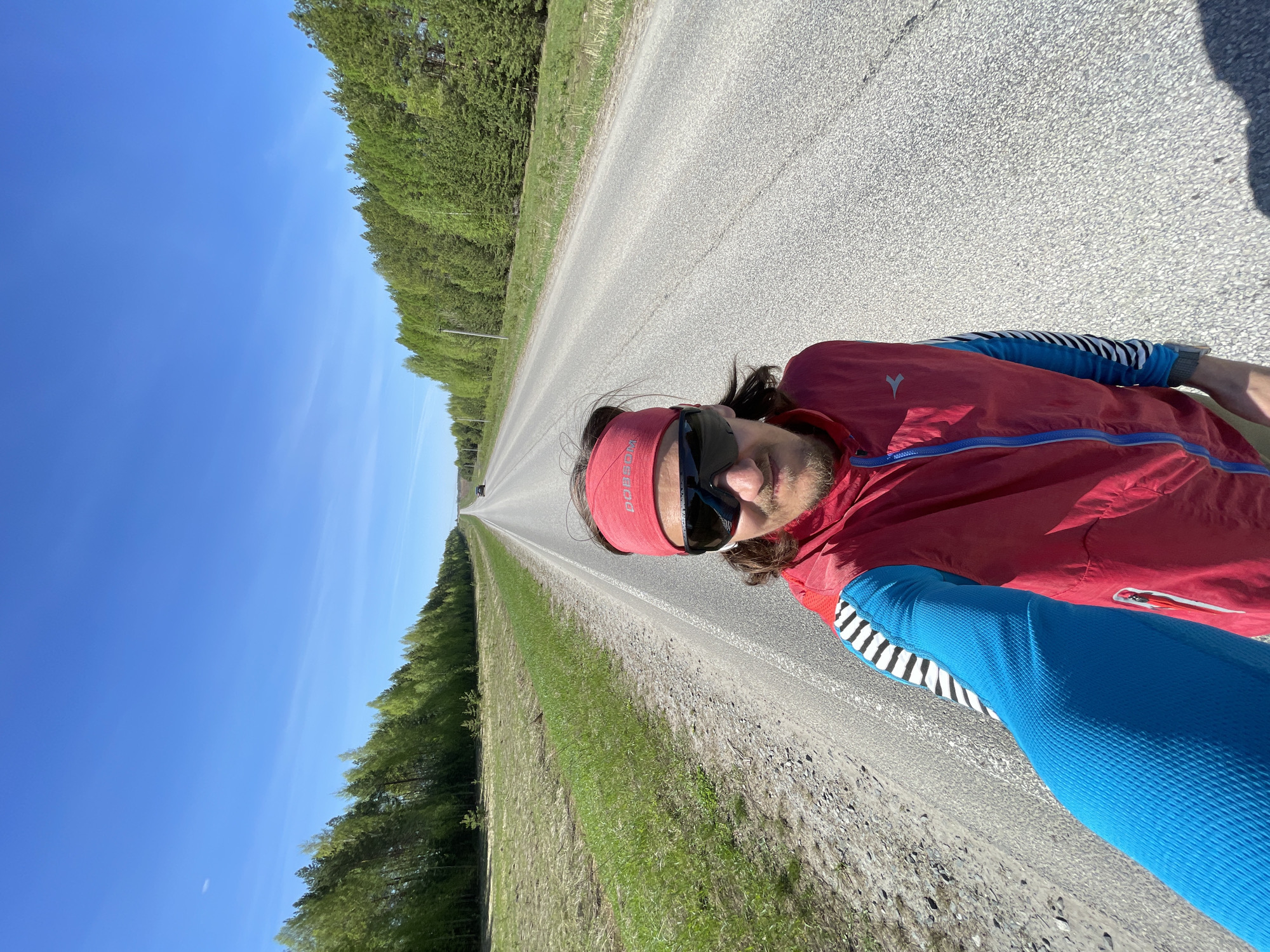 A man wearing sunglasses, a red headband with white lettering, a red and blue jacket, and blue sleeves with black and white stripes is standing on a long, straight asphalt road. He appears to be taking a selfie. The road stretches into the distance, flanked by green trees on both sides. The sky is clear and blue. The location is Перевозное, Russia, based on the EXIF data. A vehicle is visible far down the road. The terrain to the sides of the road is primarily grass.