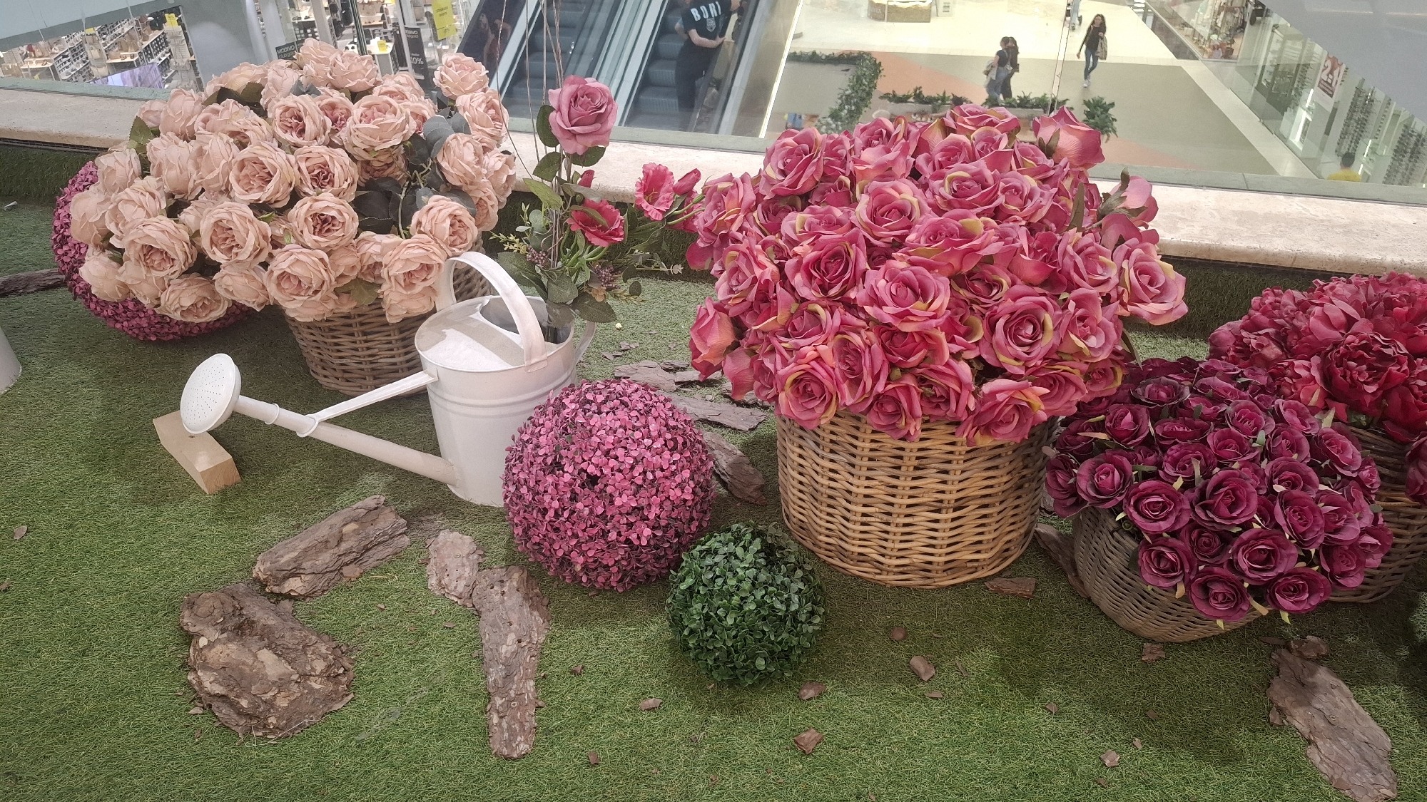 This media file captures a vibrant artificial flower display set up on a green artificial turf, likely within a shopping mall in Sofia, Bulgaria. The foreground is dominated by various arrangements of imitation flowers, predominantly roses, housed in wicker baskets.
On the left, a large basket overflows with soft pink/peach roses, while to its right, a white metal watering can with a long spout lies on its side. Nearby, a large fuchsia-colored sphere, densely packed with small artificial flowers, and a smaller green leafy sphere add to the decor. Scattered around these elements are several pieces of natural tree bark.
Towards the center and right, two more large baskets hold an abundance of artificial roses. One contains bright pink roses with yellowish undertones, and another showcases deep red/burgundy roses. Further right, a basket with larger, dark red artificial flowers, possibly peonies, completes the floral arrangement.
The background reveals a bustling indoor environment, typical of a shopping mall. People are visible, some walking on the tiled floor and others on escalators, their figures blurred. Glass railings and brightly lit store sections are also discernible. The lighting suggests it is daytime. On the t-shirt of a person on an escalator, the word "BORN" is faintly visible. The overall scene presents a decorative, static display designed to enhance the mall's aesthetics.