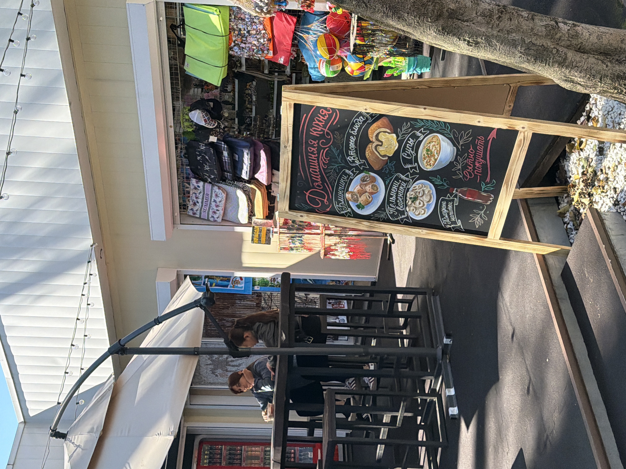 The image captures an outdoor cafe or food stall, likely in a tourist area such as Sochi, Russia, offering homemade cuisine and merchandise. The scene is brightly lit by natural sunlight, indicating daytime with clear weather.
In the foreground, two individuals are seated at a dark-colored outdoor table, beneath a large, light-colored umbrella or awning that extends from the building. One person, wearing a dark top, looks towards the left, while the other, with longer hair, appears to be looking down or conversing. The tables and chairs are made of a dark, possibly metal, material.
A prominent feature is a large, wooden-framed chalkboard menu standing upright. The menu, written in Russian with colorful chalk illustrations, advertises "Домашняя кухня" (Homemade cuisine). Specific dishes include "Блинчики" (Pancakes/Crepes), "Вторые блюда" (Main courses), "Супы" (Soups), and "Пельмени, вареники" (Pelmeni, Vareniki - types of dumplings). It also lists "Холодные напитки" (Cold drinks) and a phrase encouraging "Сытно - покушать" (Eat heartily). Illustrations show a mushroom dish, soup, dumplings, and what appears to be a bottle of Coca-Cola.
Behind the chalkboard, attached to a light-yellow building with horizontal siding, is a display of various merchandise. These include colorful bags, hats, and other items, with one bag clearly labeled "SUMMER." Other items hanging suggest beach gear or souvenirs, such as colorful balls and bright fabrics. String lights are strung along the edge of the building or awning, suggesting the area might also operate in the evenings. The ground consists of dark paving, with some white gravel and fallen leaves visible under a tree to the right, further emphasizing the outdoor setting.