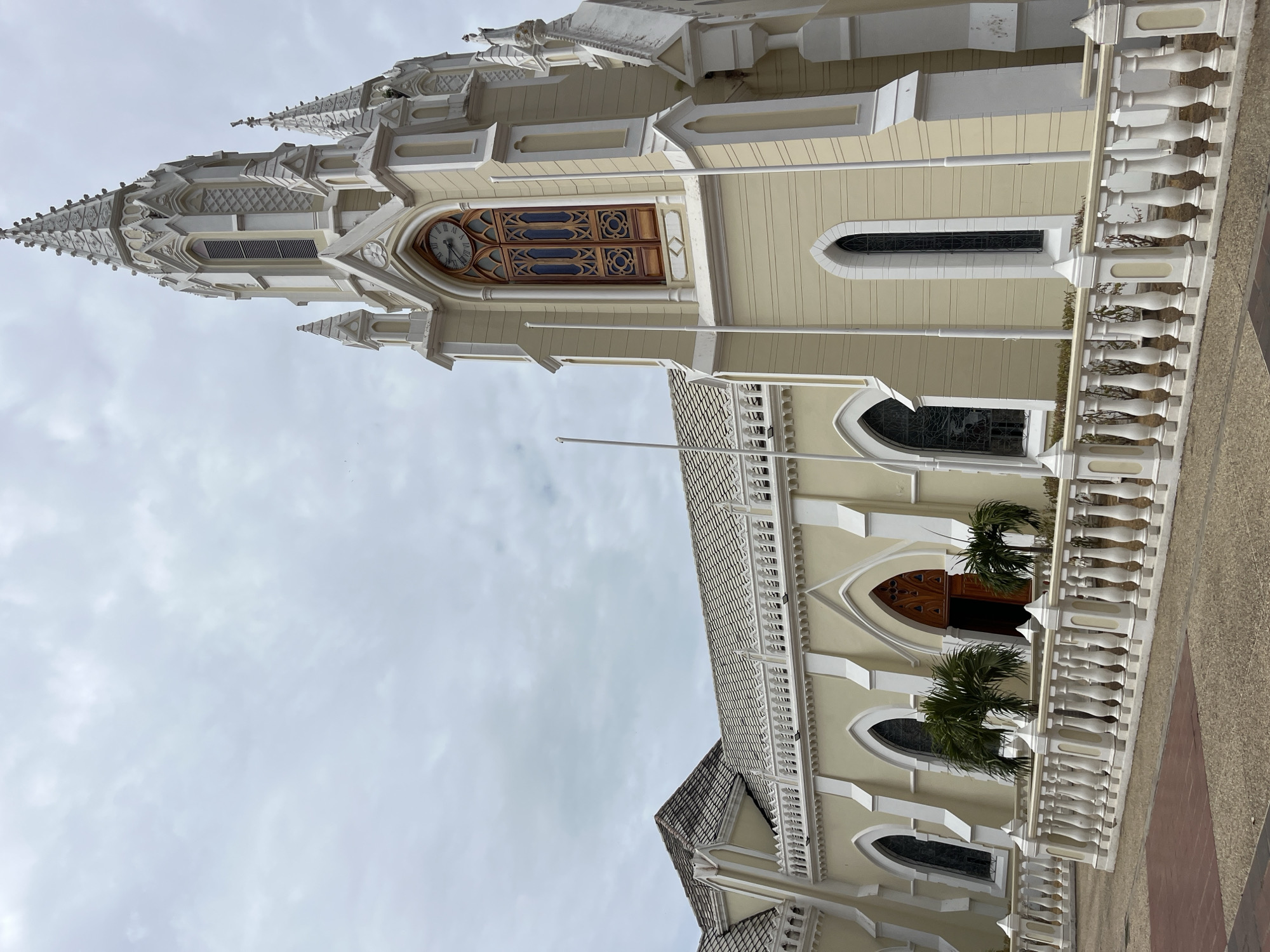 An exterior, low-angle view depicts a grand church building in El Valle del Espíritu Santo, Venezuela, under an overcast sky. The church features light cream-colored walls contrasted with white architectural details, including elaborate trim, pilasters, and a decorative balustrade running along a lower section. Multiple tall, ornate spires, characteristic of Neo-Gothic or Eclectic architecture, rise towards the top of the frame, adorned with finials and intricate patterns.
A prominent clock tower section displays a circular clock face with visible Roman numerals (I through XII), indicating a time close to noon, approximately 11:55. The building's facade is punctuated by several arched windows, some with dark panes, possibly stained glass, and others with ornate wooden latticework. Near an arched entrance on the lower facade, small palm trees add a touch of tropical greenery. The ground in the foreground and to the right appears to be a paved area. No people are visible, and no specific activity or event is taking place, focusing solely on the architectural beauty of the structure on a cloudy daytime.