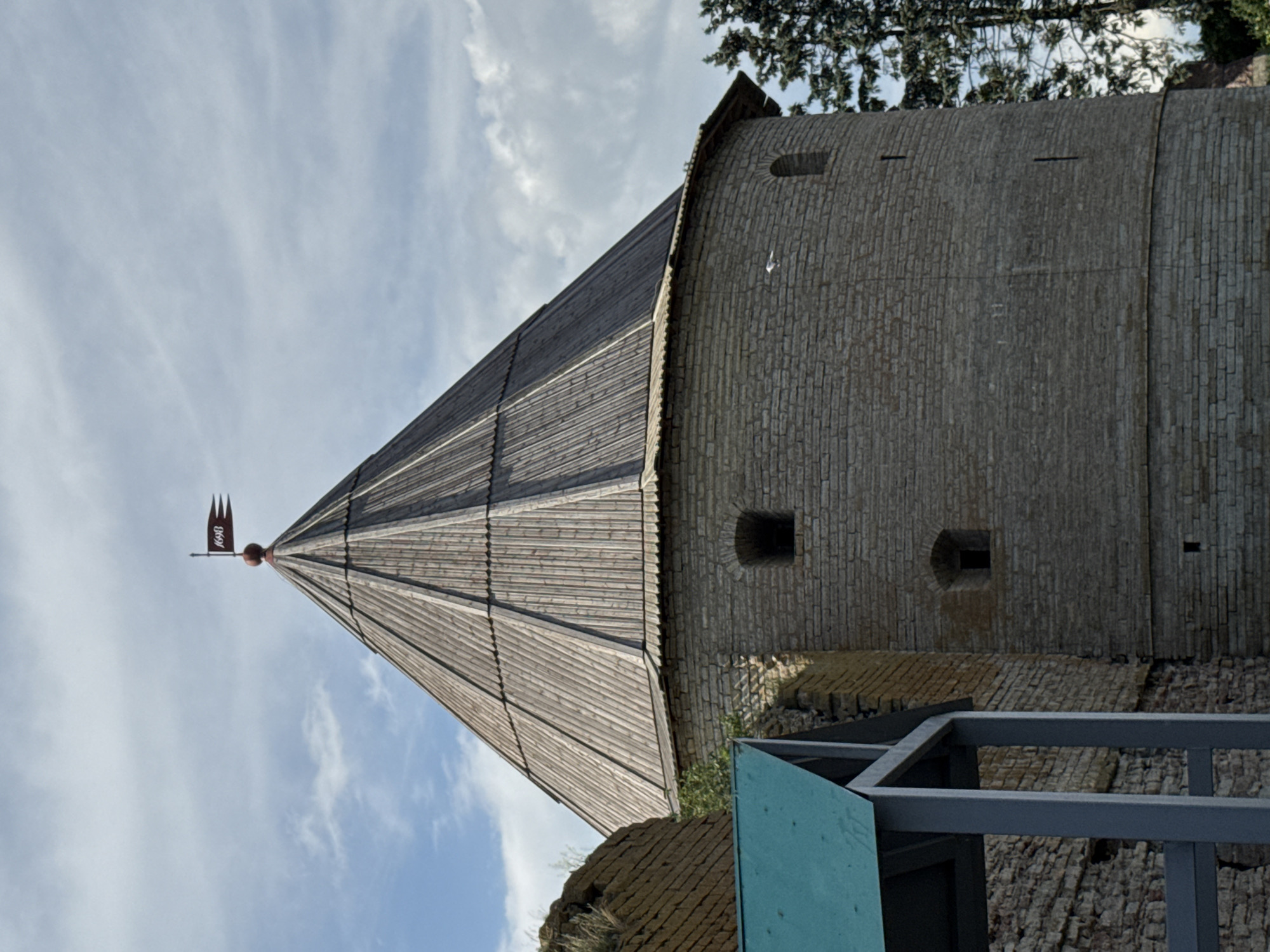 The image captures a view of a historic stone tower with a conical wooden roof, likely a part of a fortress. The tower is constructed from light-colored stone bricks, with small, dark openings visible in its walls, suggesting embrasures or windows. The wooden roof is made of planks and tapers to a point, topped with a weather vane featuring a dark red banner with some visible text.
The scene is set against a backdrop of a bright sky with scattered white clouds, indicating daytime with fair weather. Part of a green tree with dark foliage is visible at the top right of the frame, and glimpses of other stone structures, possibly ruins or foundations, are present at the bottom. A metal railing and a turquoise colored panel are also in the foreground, suggesting modern construction or maintenance work around the historic site. The overall impression is of a historical landmark being observed, with some elements of modern intervention. The orientation of the image is vertical, as if taken from a low angle looking upwards. No people are visible in the image.
The text on the banner of the weather vane is partially visible. It appears to be in Cyrillic script and reads "1640". This likely indicates a date related to the tower or the fortress. The location context of Shlisselburg, Russia, suggests this could be a part of the Oreshek Fortress.