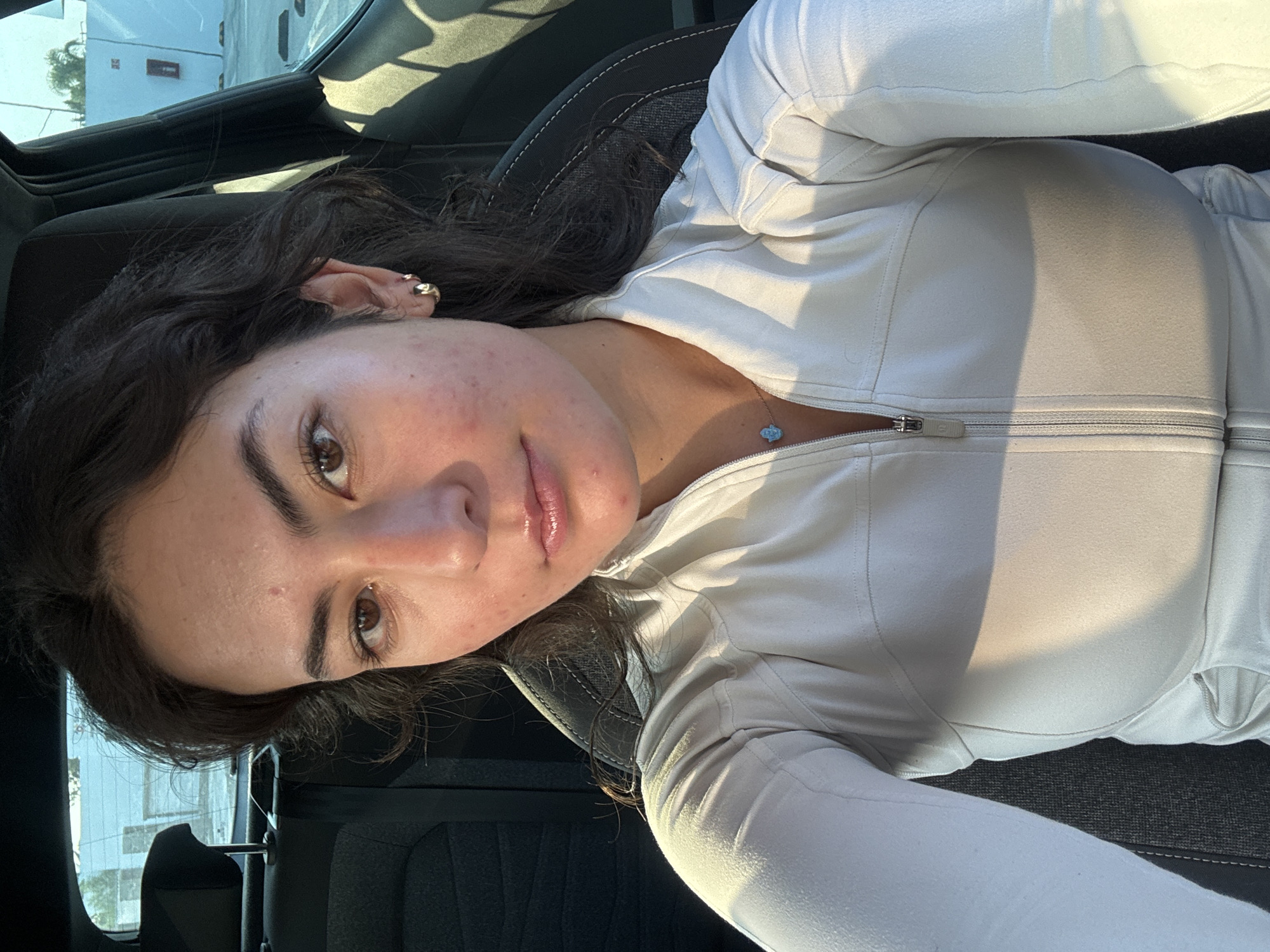 A young woman is seated in a car, possibly in San Bernardino Tlaxcalancingo, Mexico, though the exact location is not definitively identifiable from the image alone. She is wearing a light-colored zip-up jacket and has dark hair. The car's interior is visible, and a building can be seen through the window. The lighting suggests it is daytime.