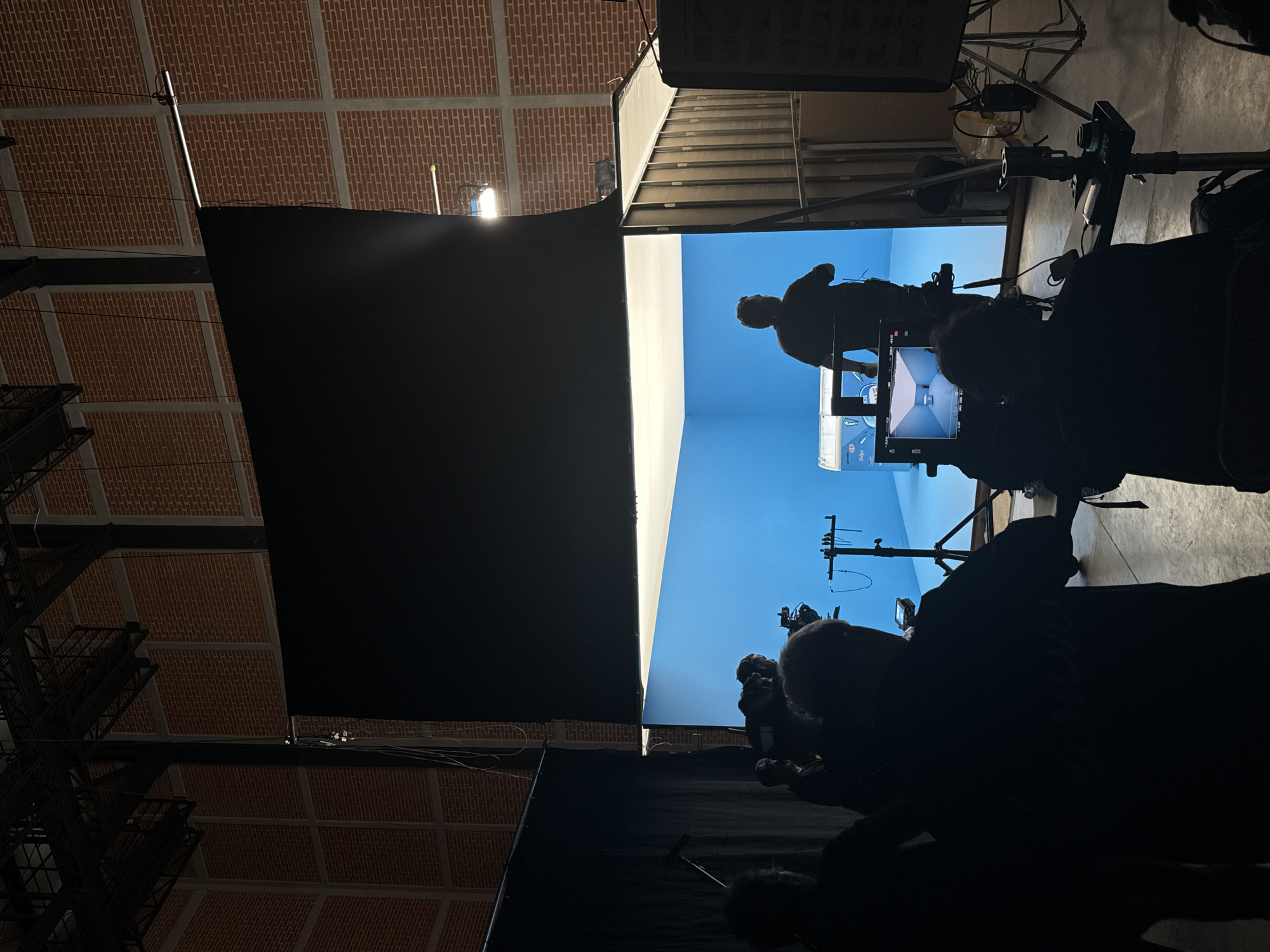 The image shows a film or video set located inside a building with a brick ceiling. A blue screen backdrop is visible. Several people are present, but they are mostly silhouetted. One person stands on a platform in front of the blue screen. Filming equipment, including cameras and monitors, are pointed towards the subject. The location is likely Tlalnepantla, Mexico.