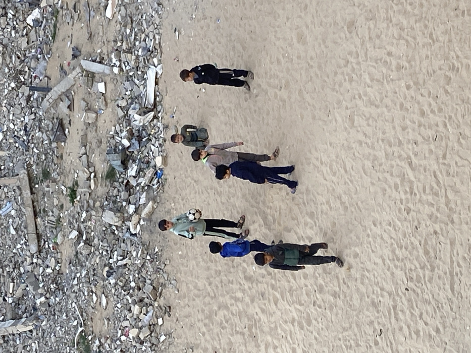 Seven young males are situated on a sandy expanse in Khan Yunis, Palestinian Territory. The background consists of a vast pile of concrete rubble, broken blocks, and debris, indicating a demolished area. The sandy ground shows visible footprints.
One boy, positioned centrally, wears a light green and black tracksuit and holds a black and white soccer ball. To his left, a boy in a royal blue tracksuit stands facing the ball-holder. To the immediate right of the boy with the ball, a boy in a light grey and beige tracksuit stands looking to the right. Further right, another boy in a black tracksuit stands with hands clasped behind his back, looking towards the center-left. A fifth boy, dressed in a dark olive green tracksuit, kneels on the sand in the midground-background. In the foreground, on the left, a boy in a dark grey and teal-green jacket with dark pants walks towards the bottom of the frame. On the right in the foreground, a boy in a dark blue tracksuit with light stripes walks towards the left. All individuals wear casual or athletic clothing.