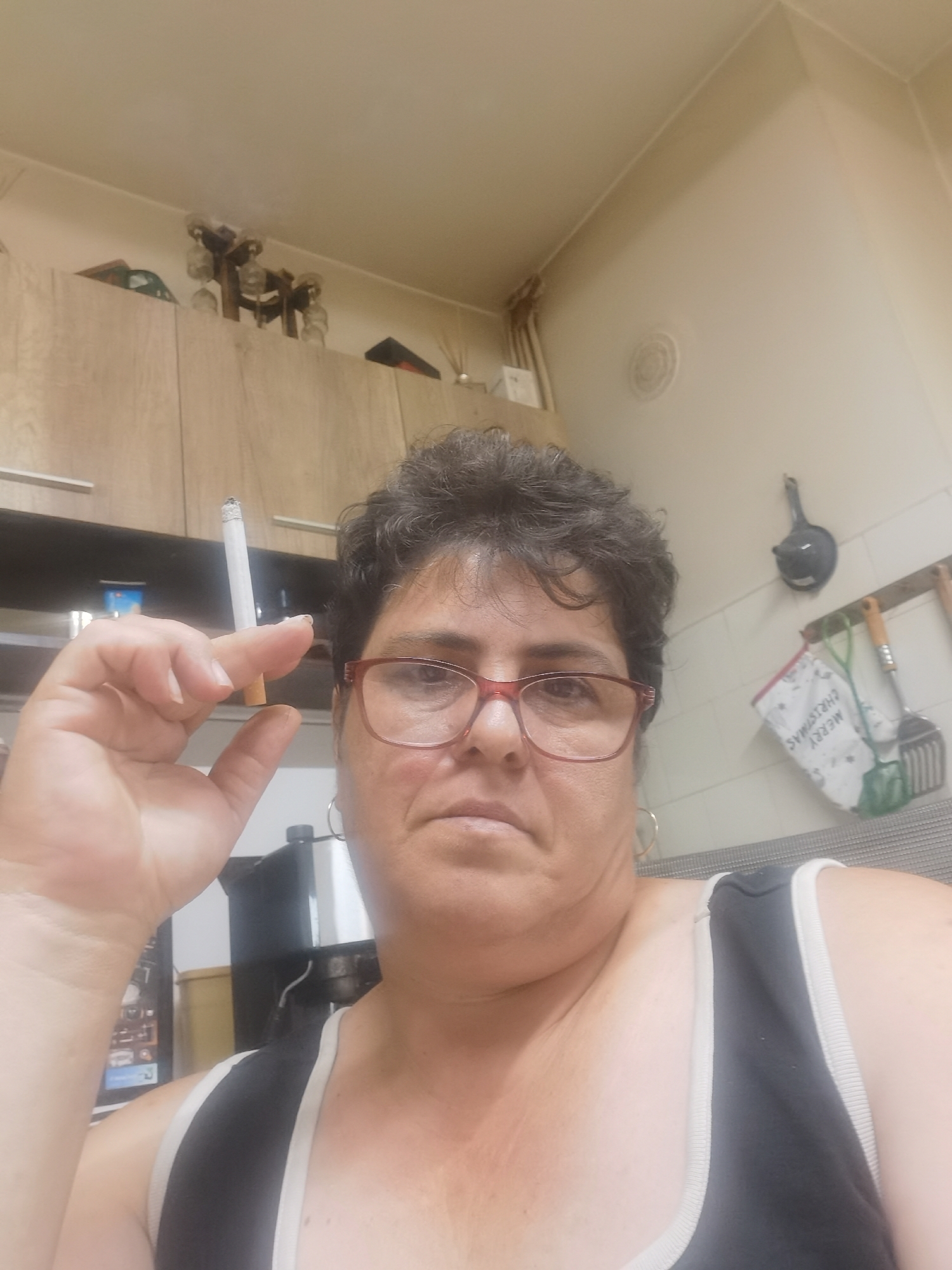 A woman is holding a cigarette between her thumb and forefinger, with the cigarette angled upwards and to her left. She is wearing glasses with red frames and a black tank top with white trim. The background appears to be a kitchen with wooden cabinets, a stove or coffee maker, and various kitchen utensils hanging on the wall. There is a decorative element on the ceiling, possibly a light fixture, and what looks like a string of lights above the cabinets. The overall lighting suggests an indoor environment, possibly during the daytime. The image was captured on a mobile phone, indicated by the angle and composition.