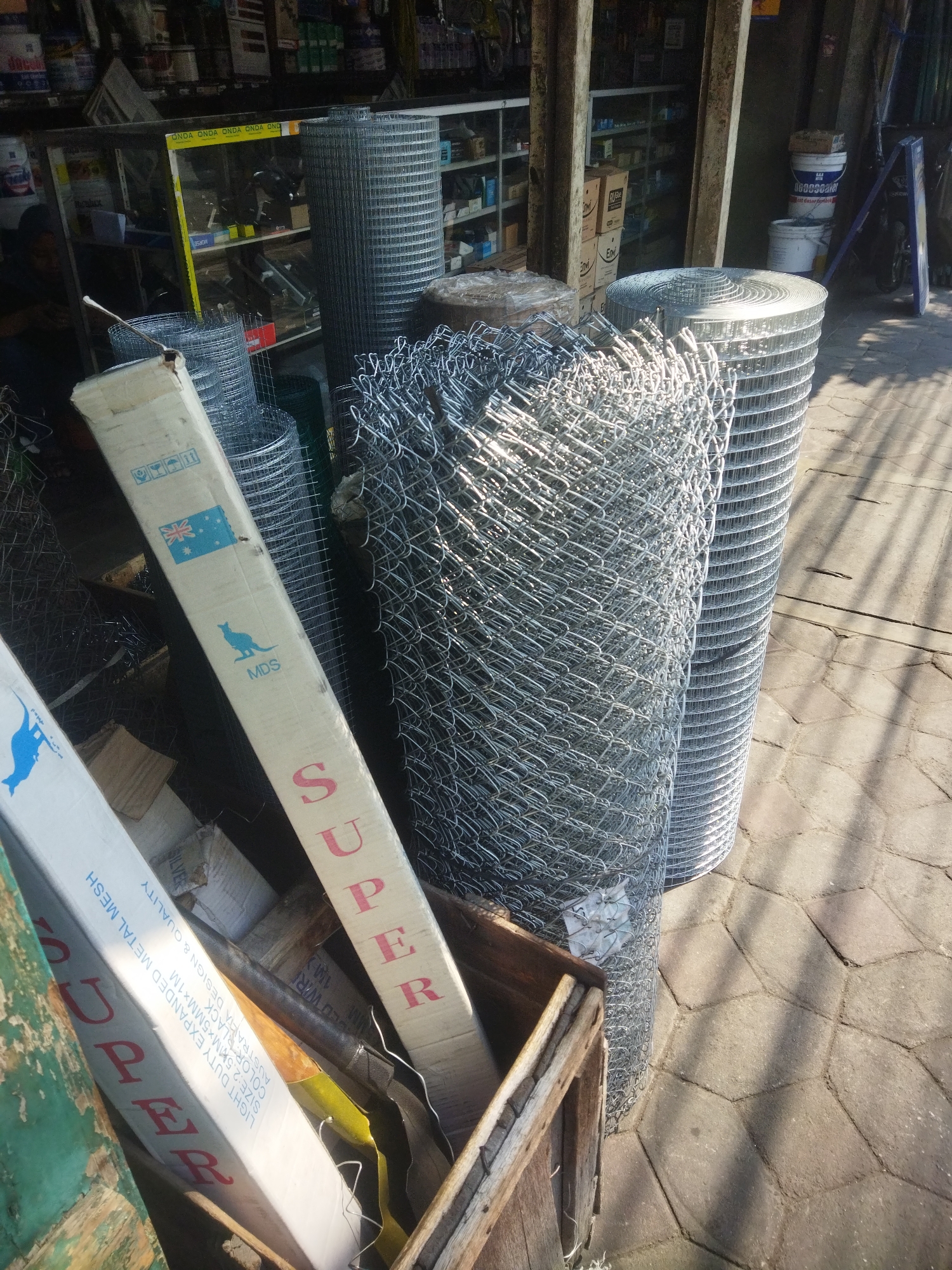 The image shows a collection of wire mesh rolls and bundled wire mesh, likely for sale at an outdoor market or hardware store in Giripurwo, Indonesia. Several large rolls of galvanized wire mesh are stacked upright, and smaller rolls are leaning against them. In the foreground, a wooden crate contains more wire mesh, along with cardboard boxes that have "SUPER" printed on them in red lettering. One box also features the Australian flag and a silhouette of a kangaroo, suggesting imported goods. The ground is paved with interlocking concrete stones, and shadows from overhead structures suggest it is daytime. The background shows shelves stocked with various items, typical of a retail environment. There are no people clearly visible in the frame.