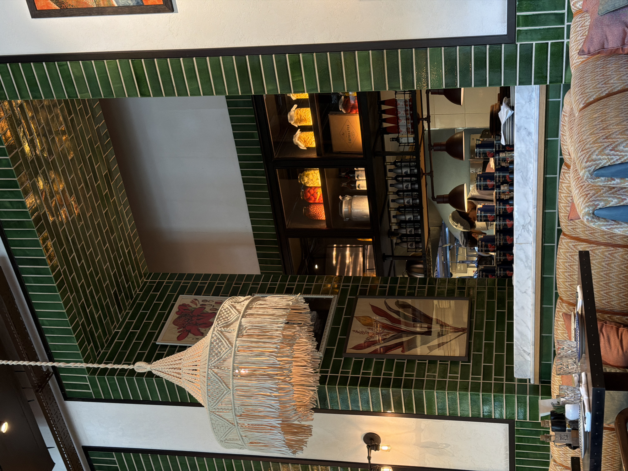 The image depicts the interior of a restaurant in Sabail Raion, Azerbaijan, with a focus on a table setting and a view into the kitchen area. In the foreground, a dark wooden table is set with two place settings, each including a white plate, silverware, and glassware. The table is positioned in front of a booth upholstered in a patterned fabric with orange and beige tones, complemented by throw pillows.
Dominating the upper left of the frame is a large, macrame hanging lamp with tassels. The walls are adorned with green subway tiles and framed artwork featuring botanical illustrations. Behind the table, a counter area is visible, leading to an open kitchen.
The kitchen area reveals shelves stocked with various jars of preserved food, bottles of wine, and cans. Three industrial-style pendant lights hang over the counter. A chef, wearing a cap and apron, is partially visible in the background, along with stacks of plates and cans on the counter. The overall ambiance suggests a well-lit, stylish dining establishment.