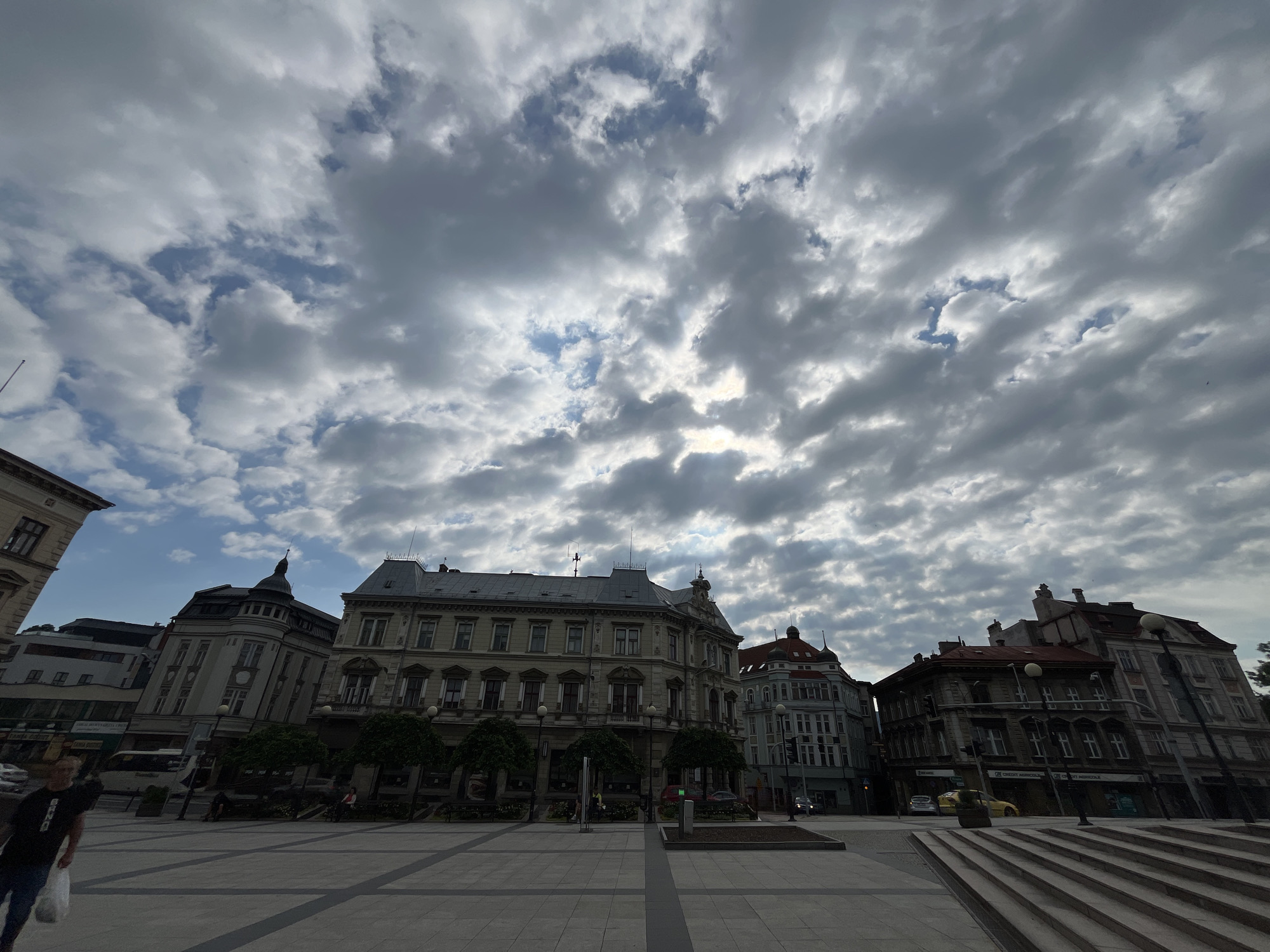 The image shows a wide-angle view of a city square under a dramatic, cloudy sky. The sky is filled with a mix of dark, ominous clouds and brighter patches, suggesting an overcast day with some sunlight filtering through.
Dominating the mid-ground are several historic buildings with ornate architecture. These buildings have multiple stories, decorative facades, and mansard roofs. Some feature arched windows, balconies, and intricate detailing. The colors of the buildings range from light gray and cream to darker tones, with some accents of red on a rooftop.
In the foreground, a paved plaza is visible. The paving stones are laid out in a geometric pattern. A set of wide, stone stairs is prominent on the right side of the image, leading up towards the buildings.
There are a few people present in the scene. On the left side, a person in a dark t-shirt and jeans is walking away from the camera, carrying a white bag. On the plaza, several other individuals are seated on benches or walking in the distance, appearing as small figures. Trees with dark green foliage are planted along the front of some of the buildings, providing a touch of nature to the urban landscape.
A bus is visible in the background, and cars are present on the street beyond the plaza. Streetlights are scattered throughout the square. The overall impression is one of a historical European city square on a day with dynamic weather.