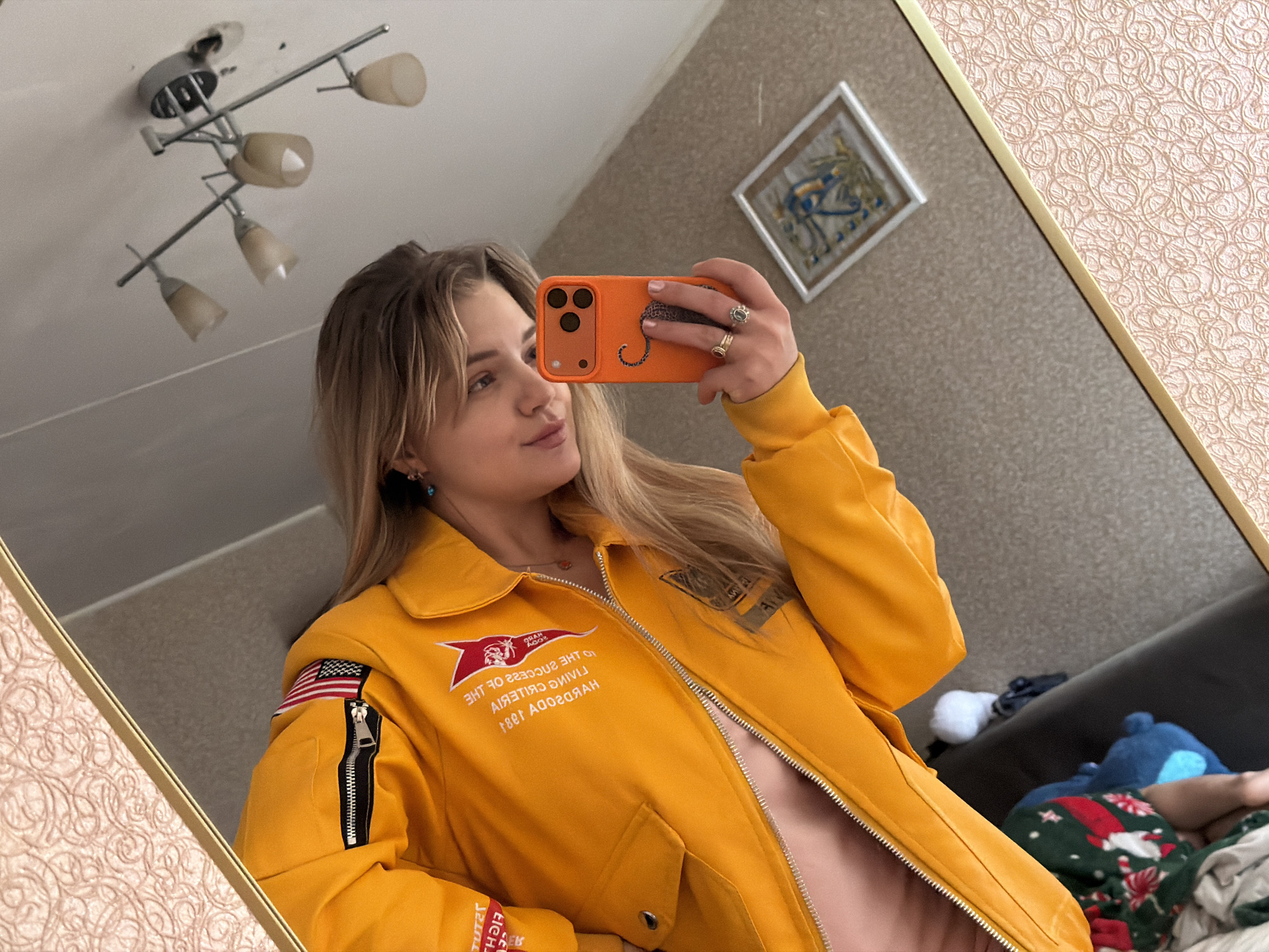A woman is taking a mirror selfie. She has blonde hair and is wearing a yellow jacket. She is holding an orange smartphone. The background includes a ceiling light fixture, a framed picture, wallpaper with a floral pattern, and a glimpse of a sofa with items on it. The location is likely inside a home or apartment, possibly in Naberezhnye Chelny, Russia.