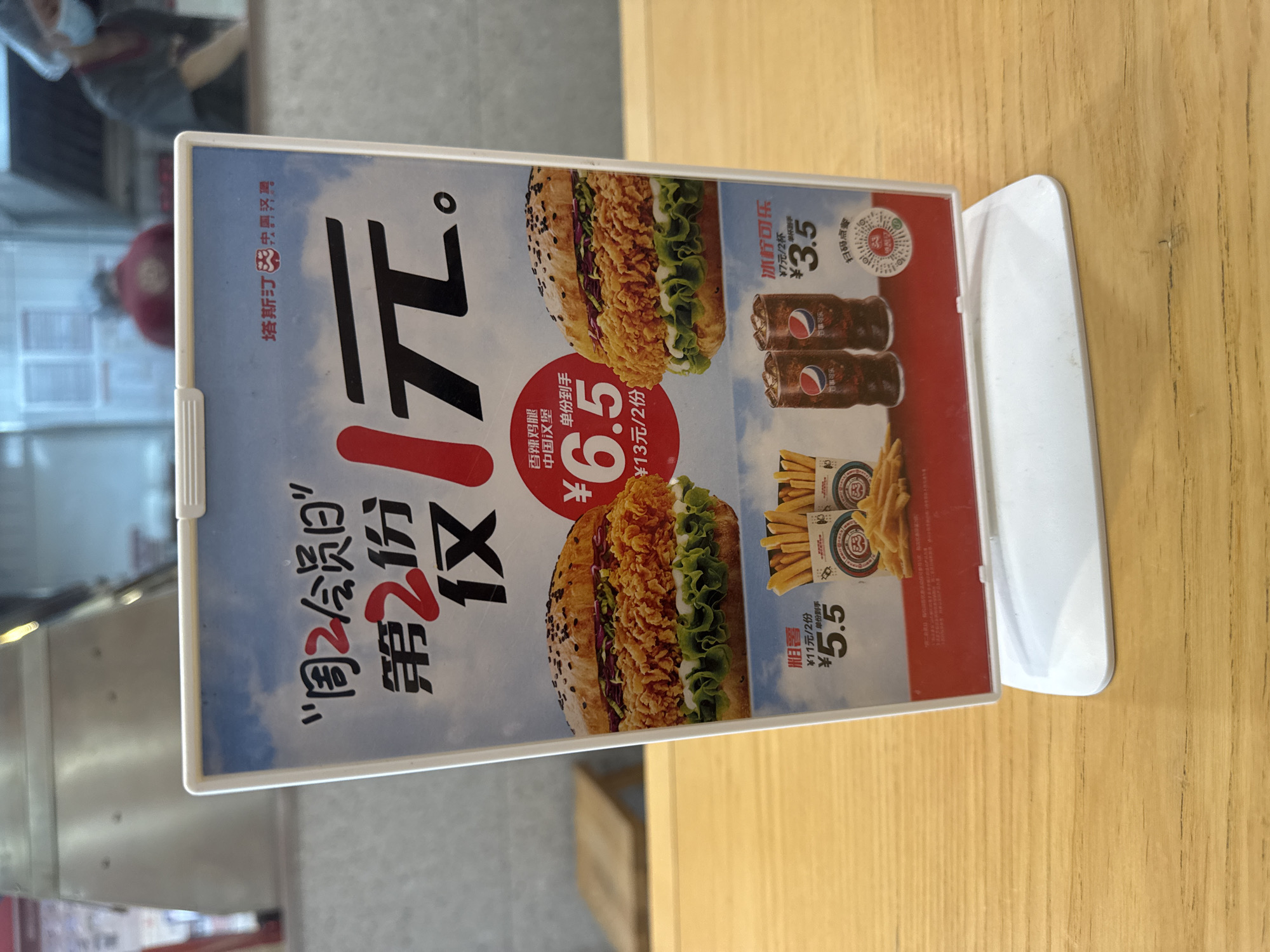 A promotional menu sign is displayed on a wooden table in what appears to be a fast-food establishment. The sign features an advertisement for a "Member Day" special, offering the second item for 1 yuan. The menu prominently displays images of two large burgers and two cups of Pepsi. Below the burgers, there are offers for fries and Pepsi. The sign is white and blue with red accents and Chinese text. In the background, a blurred view of employees in uniform and a food preparation area is visible. The overall scene suggests a casual dining environment.