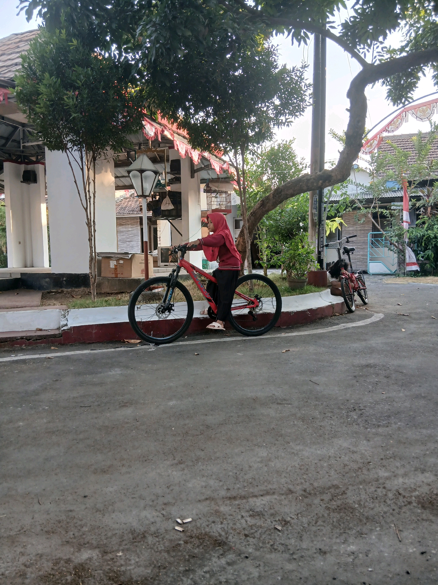 In Jepangpakis, Indonesia, a young person wearing a red hijab and a red jacket is standing next to a red bicycle. They are on a paved surface, likely a road or sidewalk, with a curb to their left. The background features a white building with a tiled roof and red and white decorations, suggesting a festive or national occasion. There are trees and foliage surrounding the area, and another red bicycle is visible further down the path. The lighting indicates it is daytime, with sunlight filtering through the trees.