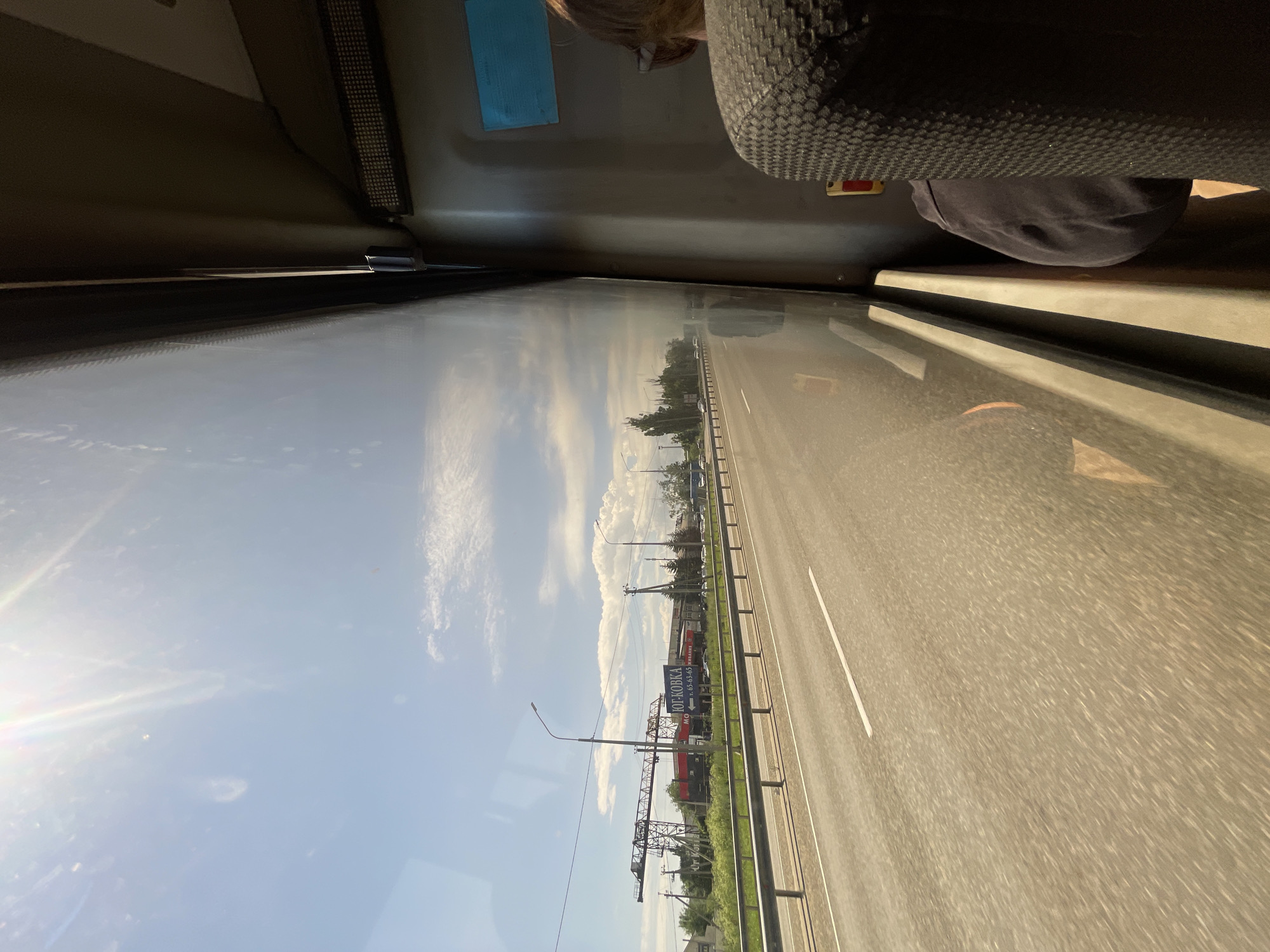 The image captures a view from inside a vehicle, likely a bus or train, looking out a window. The perspective is unusual, as the image is rotated approximately 90 degrees clockwise.
In the foreground, the interior of the vehicle is visible. A dark-colored seat with a textured surface is on the right side, and a portion of a person's dark clothing is also visible. Above, a light-colored panel lines the interior of the vehicle.
The main scene observed through the window is a highway with multiple lanes. The road surface is asphalt, and white lane markings are clearly visible. On the left side of the highway, there is a clear blue sky with scattered white clouds, suggesting fair weather. The sun is shining, casting lens flare across the left side of the frame, indicating it's likely daytime, possibly mid-morning or afternoon.
Along the right side of the road, there are green trees and vegetation. In the distance, there are several structures and signs. One prominent blue sign with white text, which appears to be in Cyrillic, reads "ЮГ-КОВКА" (YUG-KOVKA) and includes a phone number. There are also some industrial-looking structures, possibly cranes or scaffolding, visible in the background. The scene suggests a rural or suburban setting along a major road. The overall atmosphere is one of travel and transit.