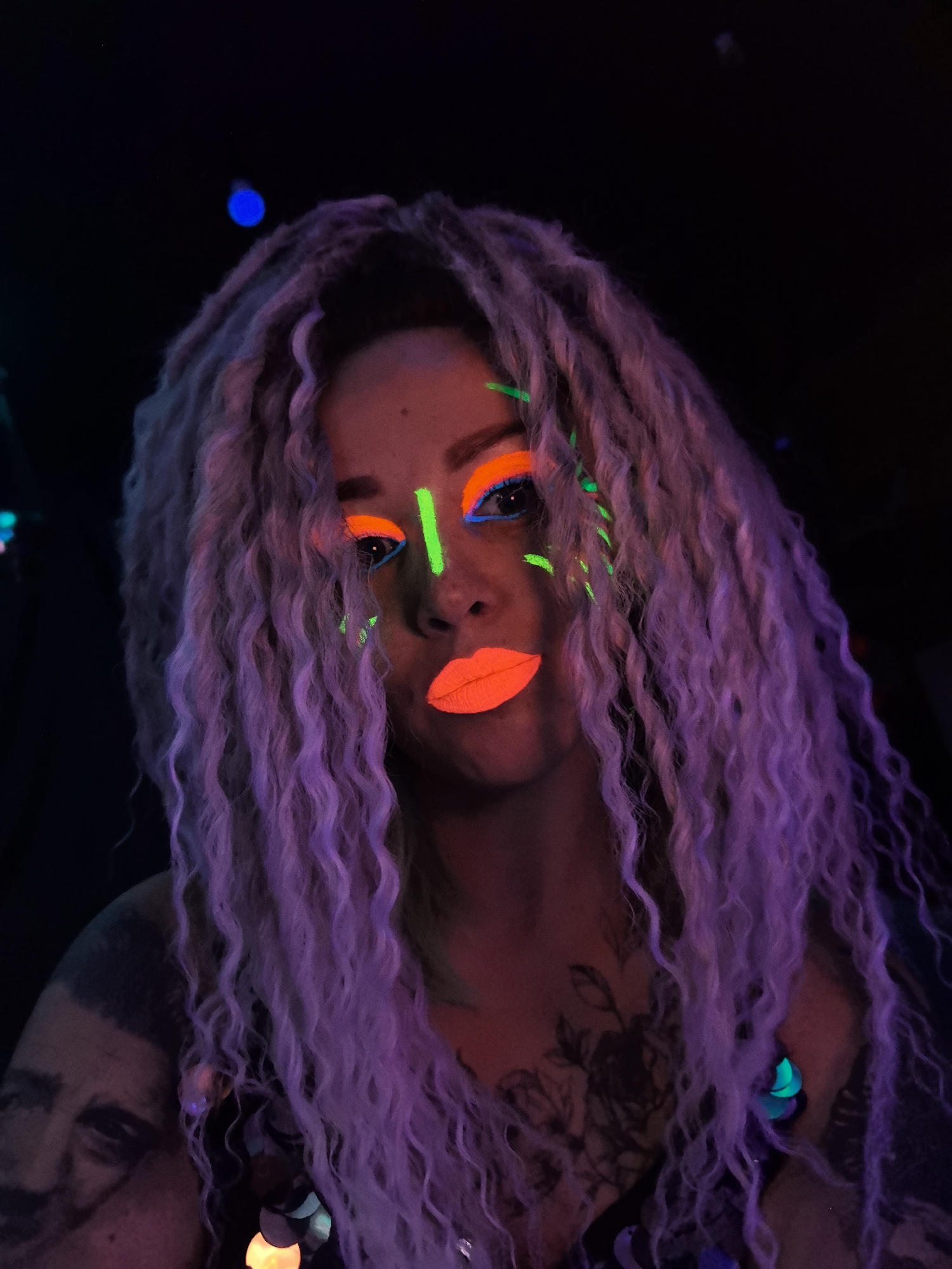 A person with long, light-colored wavy hair and vibrant neon makeup poses for a close-up portrait. The person's eyes are adorned with bright orange eyeshadow and green accents, and their lips are painted a striking orange. A green stripe is drawn on their nose. The scene is illuminated by a blacklight, giving the hair a lavender hue. The person has tattoos on their shoulder and chest. The background is dark. There is no location information in the city of Noviy, Russia.