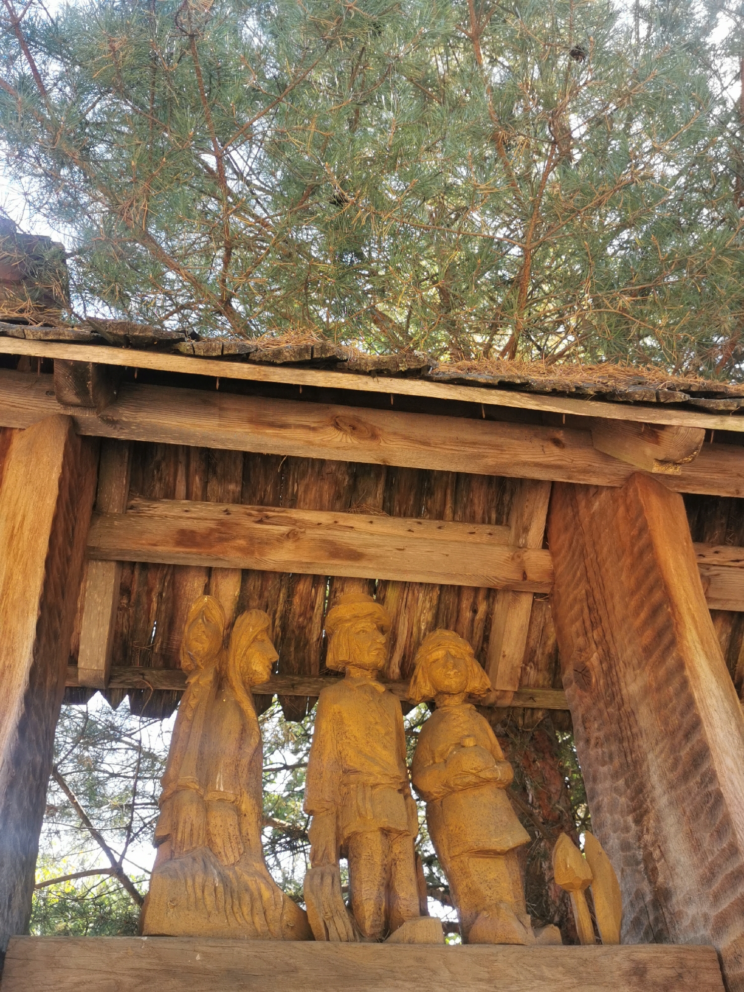 The image depicts a wooden structure with carved wooden figures underneath. The structure appears to be a rustic shelter or pavilion, with a roof made of wooden shingles and beams. The wooden figures are carved in a folk art style, depicting individuals with simple clothing and hairstyles. The figures are arranged in a row, with the leftmost figure appearing to be a woman, followed by two children. The environment suggests an outdoor setting, possibly a park or a village square, with trees and foliage visible in the background. The lighting indicates it is daytime, with sunlight filtering through the trees. The overall impression is one of a handmade, decorative installation, likely part of a local cultural attraction. The specific location in Шчарчоўскі сельскі Савет, Belarus, suggests a rural or semi-rural setting within the country.