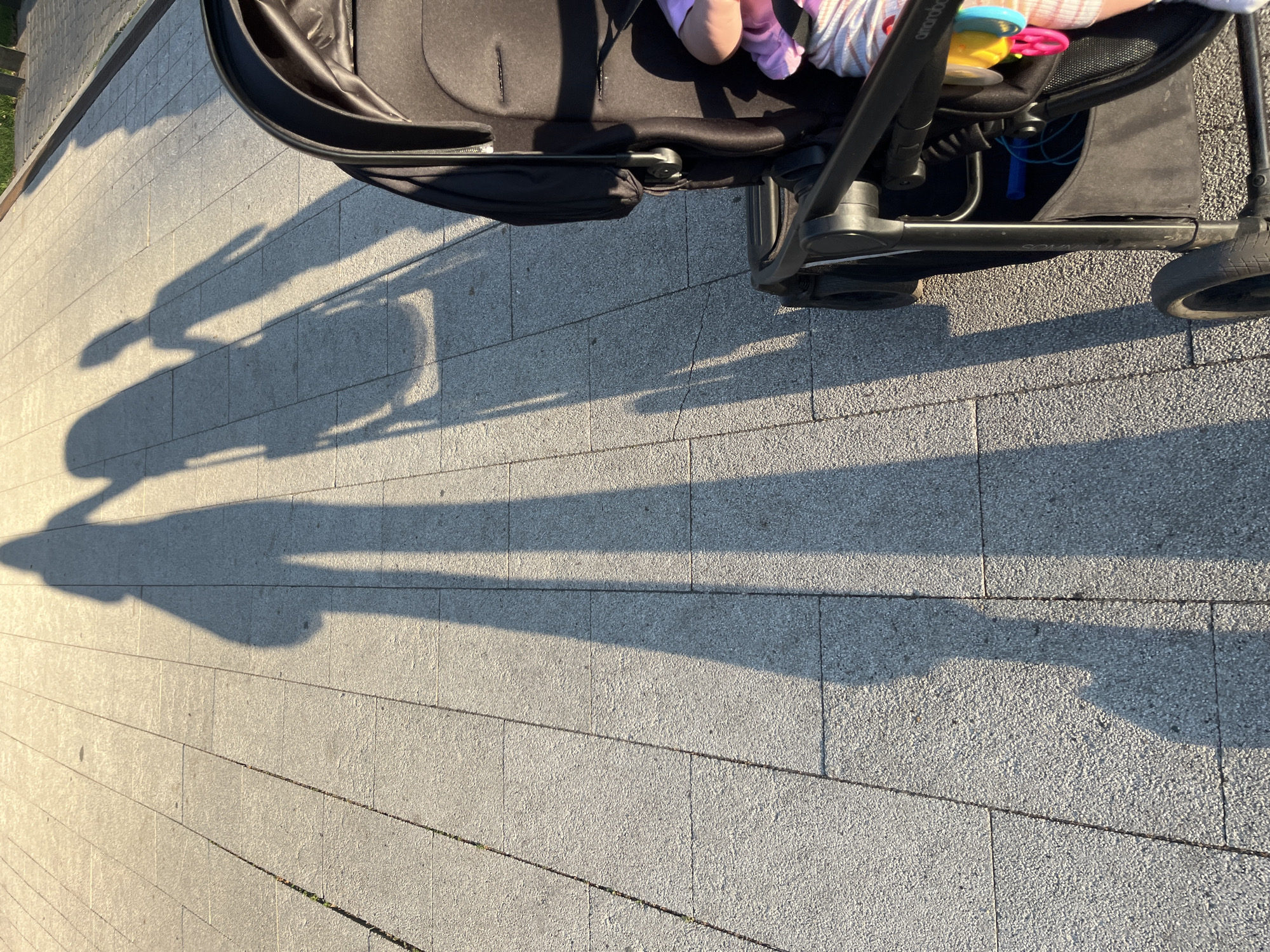 A long shadow of a person holding hands with a child is cast on a stone-paved walkway. The shadows stretch from left to right, indicating the sun is low in the sky, likely during golden hour. To the right of the walkway, a portion of a black baby stroller is visible, with a baby's arm in a striped outfit peeking out from within. The stroller has toys attached to it. The walkway appears to be in an outdoor public space. The location in Novaya Usman', Russia is a detail not deducible from the image itself.