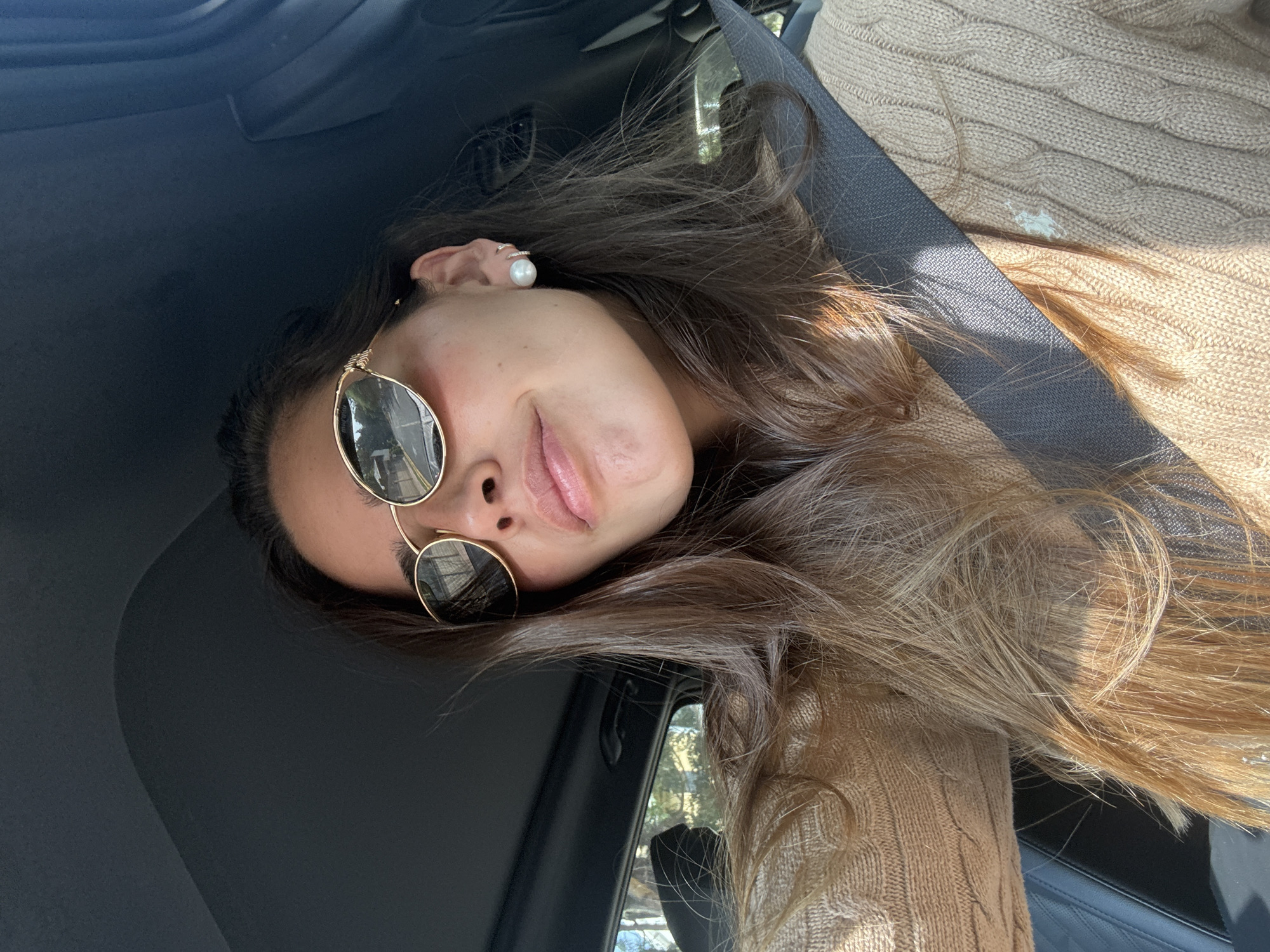 A close-up, tilted portrait captures a person, likely female, inside the dark interior of a car. The subject has long, flowing brown hair with lighter, possibly sun-lightened streaks, that extends across the right side of the frame. They wear round, gold-rimmed sunglasses, which reflect bright light and outdoor greenery, suggesting a sunny day. A large, white pearl earring and a small ring are visible on their ear. The person's lips are slightly parted in a relaxed, subtle smile, conveying a sense of calm or contentment.
They are dressed in a light brown or beige cable-knit sweater. A dark grey seatbelt stretches diagonally across their chest. The background shows the car's dark headliner and a side window reflecting trees and foliage, indicating an outdoor setting. The bright illumination points to daytime. The subject appears to be reclining or leaning back, possibly taking a selfie. There is no visible text. The provided context places the scene potentially in San Bernardino Tlaxcalancingo, Mexico.