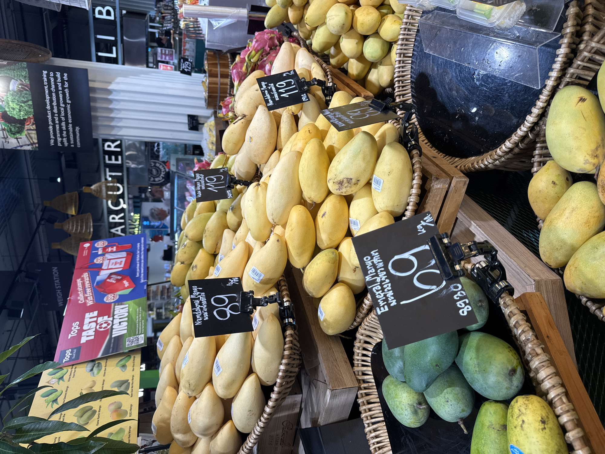 This is a medium shot, eye-level, outdoor photograph taken in the city of Baan Chaweng, Thailand. The scene is a fruit stand, likely within a market or grocery store. The foreground is dominated by a large display of yellow mangoes, arranged in woven baskets. Several price signs are visible, one indicating "Nam Dok Mai Mango" with a price of "89". Another sign displays "Emerald Mango" with a price of "69". In the background, more mangoes are visible, along with a glimpse of dragon fruit and other produce. Signs with promotional information and branding, such as "Tops" and "WEIGH STATION," are also present. The overall lighting suggests a well-lit environment, possibly with artificial lighting from the store. No people are clearly visible in the frame.