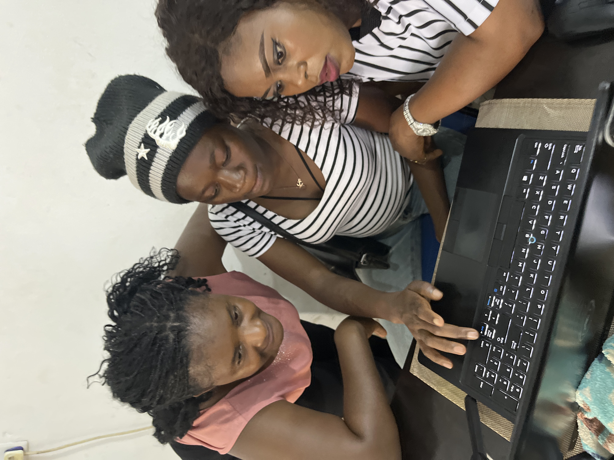 Three women are gathered around a laptop on a table. The woman on the left, wearing a pink shirt, leans in to look at the screen, her expression one of focused attention. She has braided hair. The woman in the middle wears a black and white striped shirt and a black beanie with a white star and logo. She is also looking at the laptop, with her hand resting on the keyboard. The woman on the right, also in a black and white striped shirt, looks intently at the screen, her makeup visible. She wears a watch on her left wrist. The laptop is open, displaying a keyboard with illuminated keys. The scene takes place indoors, with a plain white wall in the background. The table appears to be dark wood, and a placemat is visible beneath the laptop. The overall impression is one of collaboration or shared activity involving the laptop.