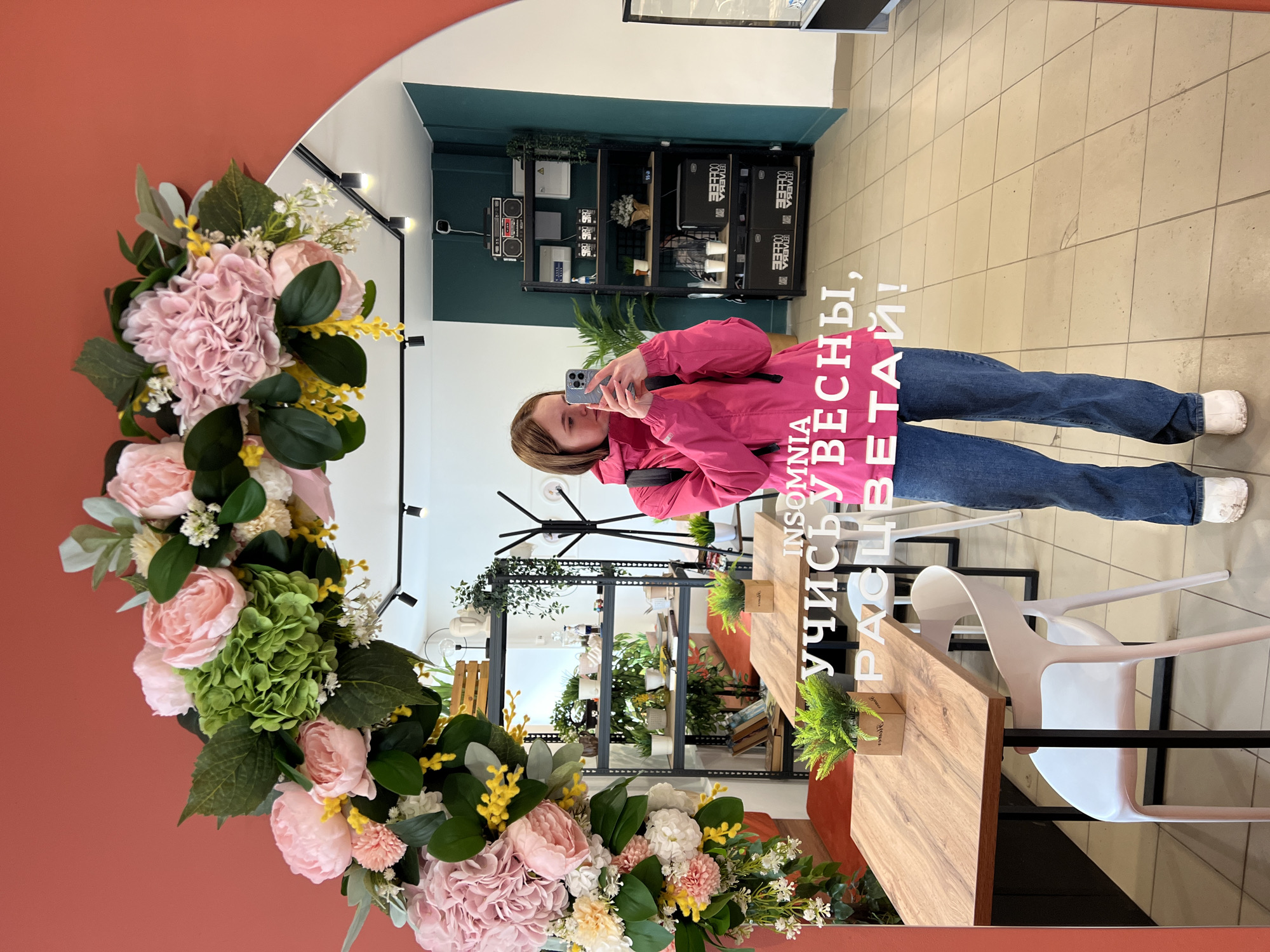 A photograph taken in Chaikovsky, Russia, shows a woman reflected in a mirror, taking a selfie. The mirror is framed by a floral arrangement, predominantly pink and white. The reflection shows the woman wearing a pink jacket and jeans. Behind her, the interior of a space with shelving units displaying plants and objects is visible. There are also tables and chairs. Text is superimposed on the image. The floor has a tiled pattern.