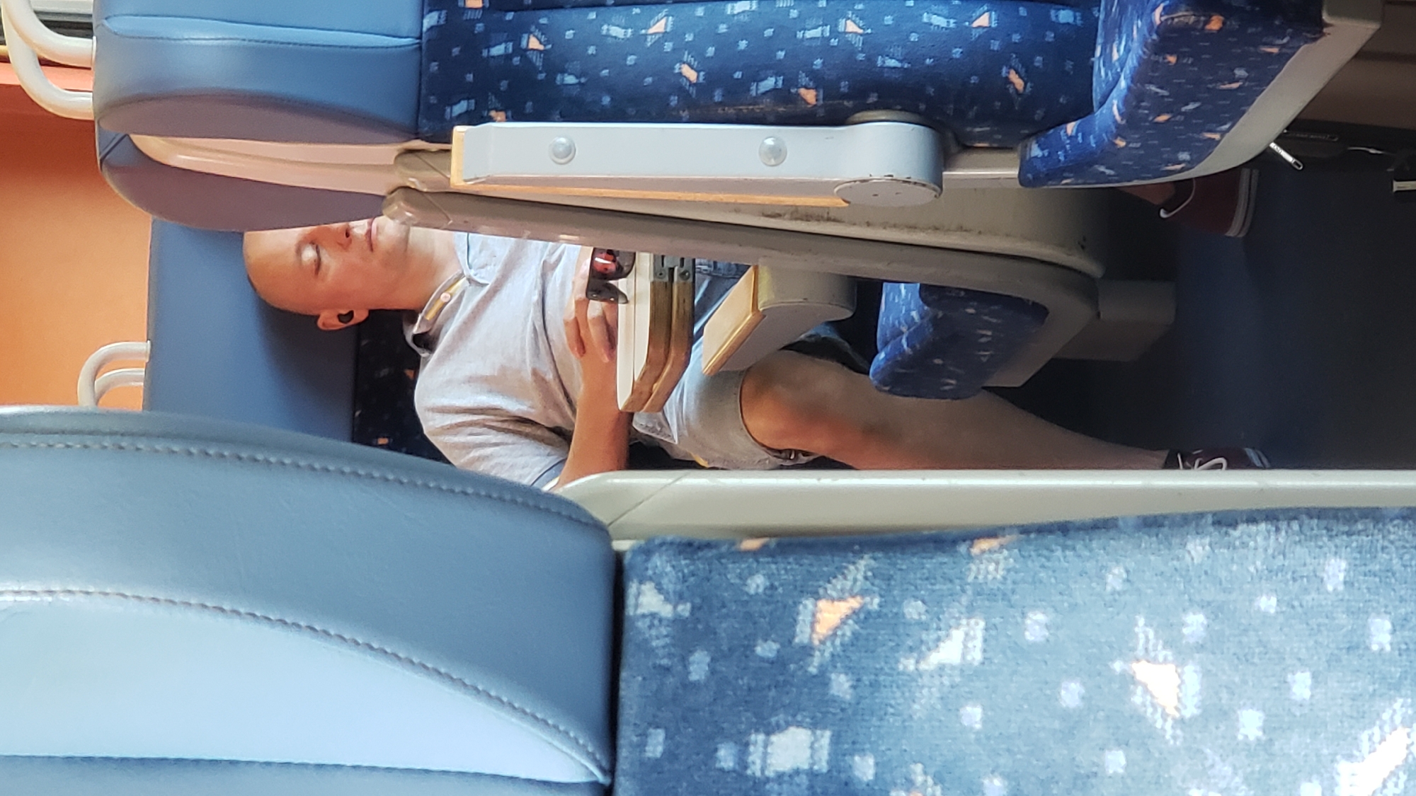 A bald man is seated on a train, sleeping with his eyes closed. He is wearing a light grey polo shirt and grey shorts, with a pair of black earbuds in his ears. A pair of sunglasses rests on a small foldable table in front of him. The seats are upholstered in blue fabric with a repeating pattern. The background shows the interior of a train car. There is no information available about the city of Považská Bystrica, Slovakia in relation to this image.