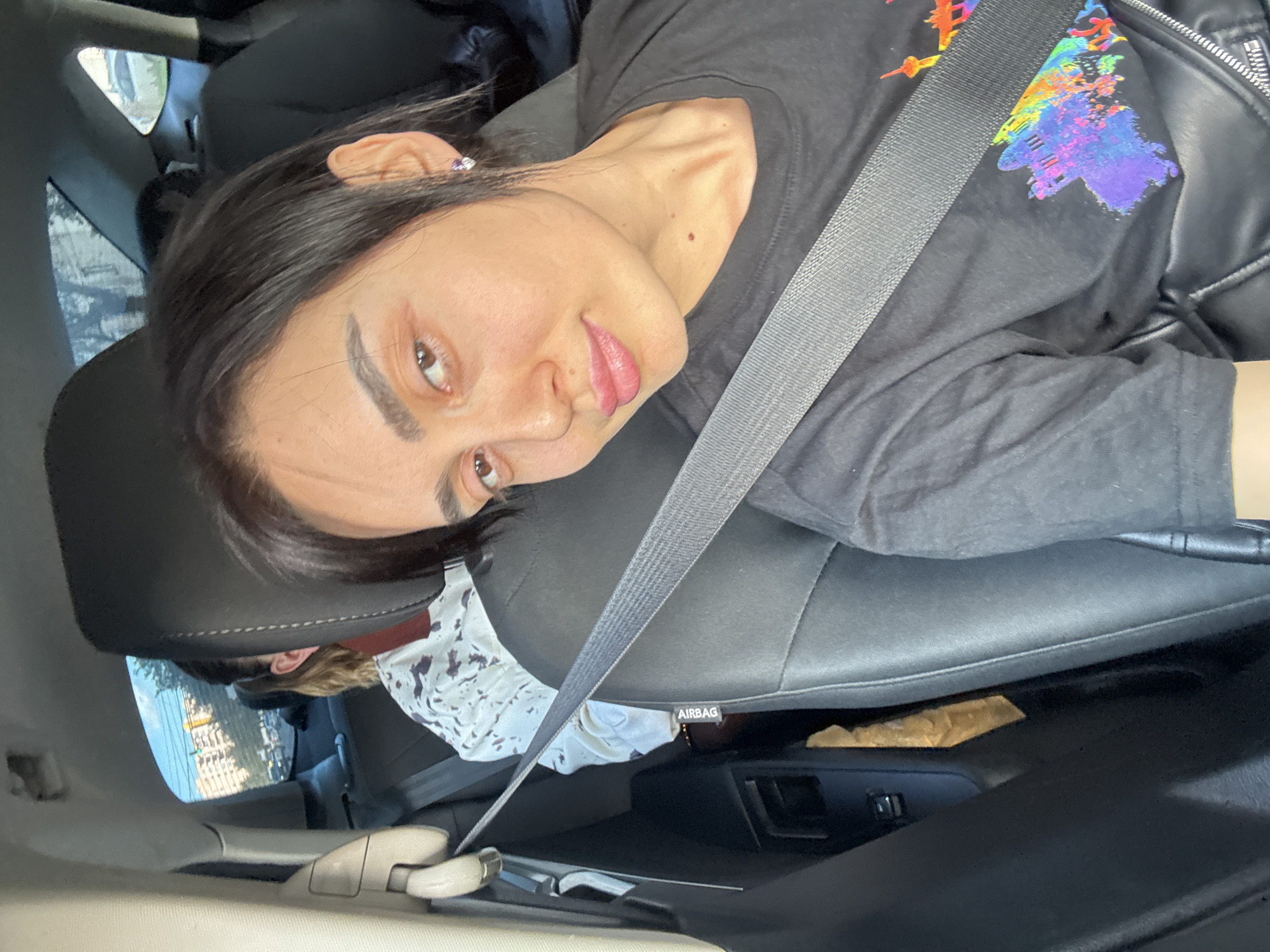 A woman with dark hair and fair skin, wearing a black t-shirt and a black leather jacket, sits in a car and looks towards the camera. She is wearing a seatbelt. Another person, with light-colored hair and wearing a patterned top, is visible in the back seat. Outside the car window, buildings and a street scene are visible, appearing to be in Tbilisi, Georgia. The interior of the car is mostly black.