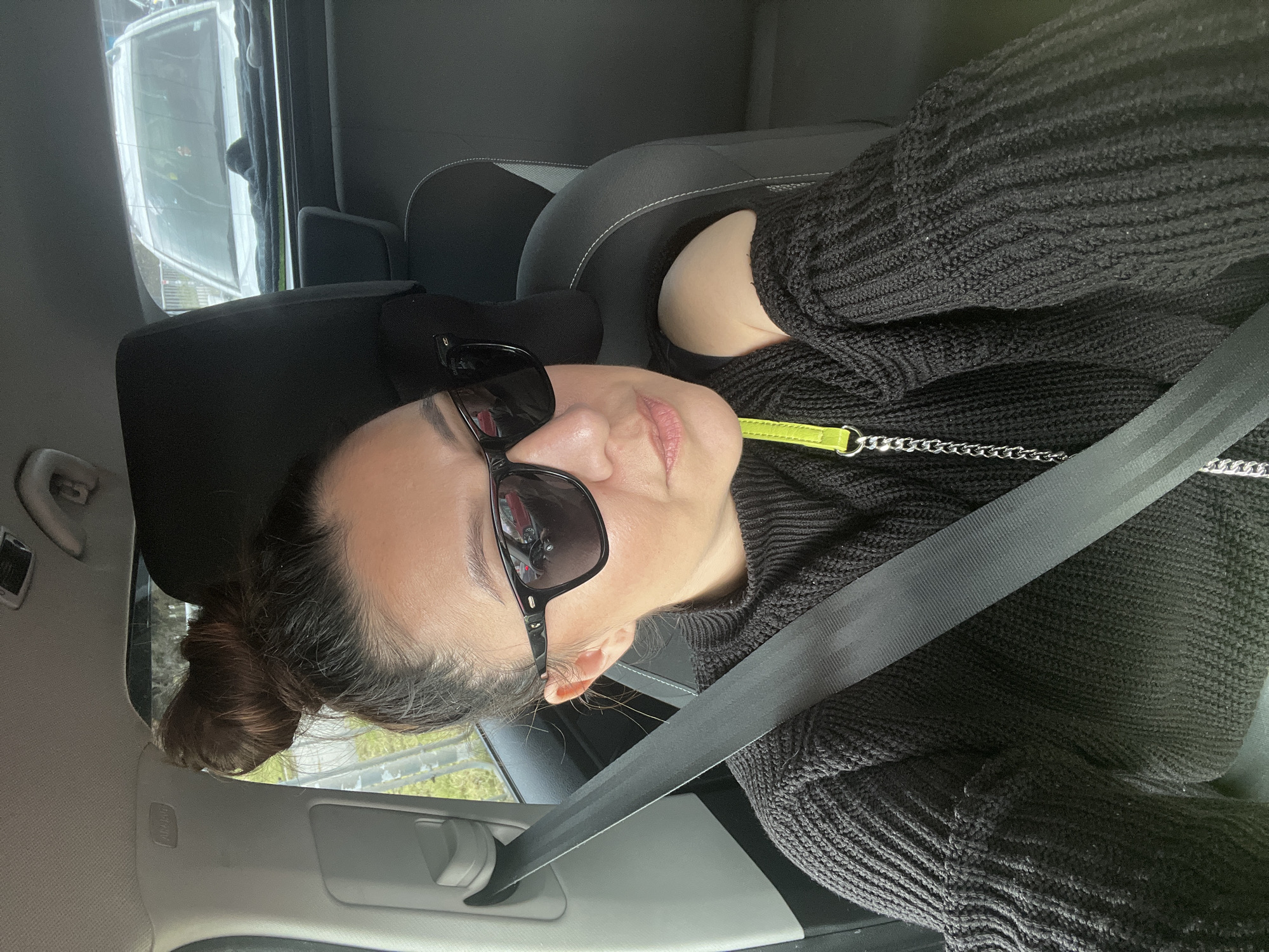 A woman is in a car, wearing sunglasses and a black sweater. She is wearing a seatbelt. The location may be Varna, Bulgaria, but there is no context in the image to support this claim. The background shows a view of the outside through the car window.