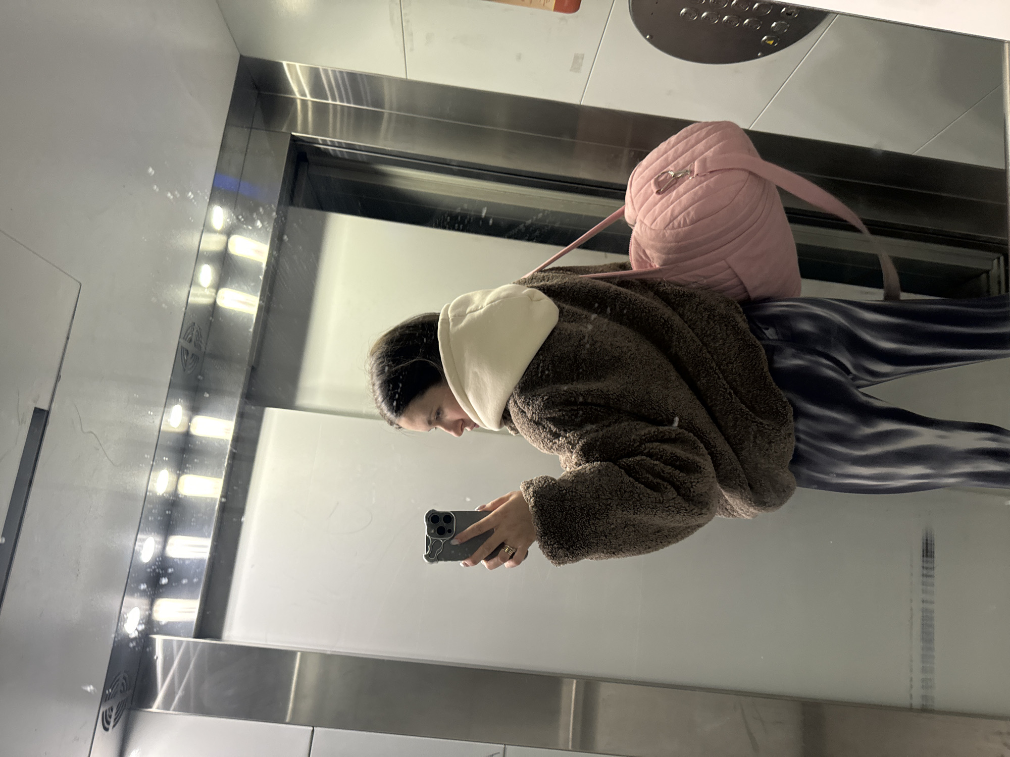 The image, taken in Odintsovo, Russia, captures a young woman taking a mirror selfie inside an elevator. The photo is oriented ninety degrees clockwise, showing the woman's reflection from the waist up. She has dark hair and is wearing a light-colored hooded top under a brown, fuzzy jacket or coat. She holds a dark-colored smartphone with multiple cameras in her right hand, seemingly focused on its screen to capture the reflection. A pink, quilted backpack with a single strap is visible on her back, suggesting she might be carrying belongings. Her lower body, partly visible, shows dark blue or black patterned pants with an abstract, wavy design.
The setting is a modern elevator interior with sleek metallic panels and light-colored walls or ceiling, which are highly reflective. Several bright, vertical strip lights are visible along the metallic frame, reflecting prominently. In the upper left corner of the reflection (if the image were rotated correctly), a circular metallic elevator control panel with multiple buttons is partially visible. No discernible text is visible on the control panel or elsewhere within the frame. The time of day and weather are not discernible due to the indoor, artificial lighting. The woman's expression is not clearly visible, but she appears to be concentrating on her phone.