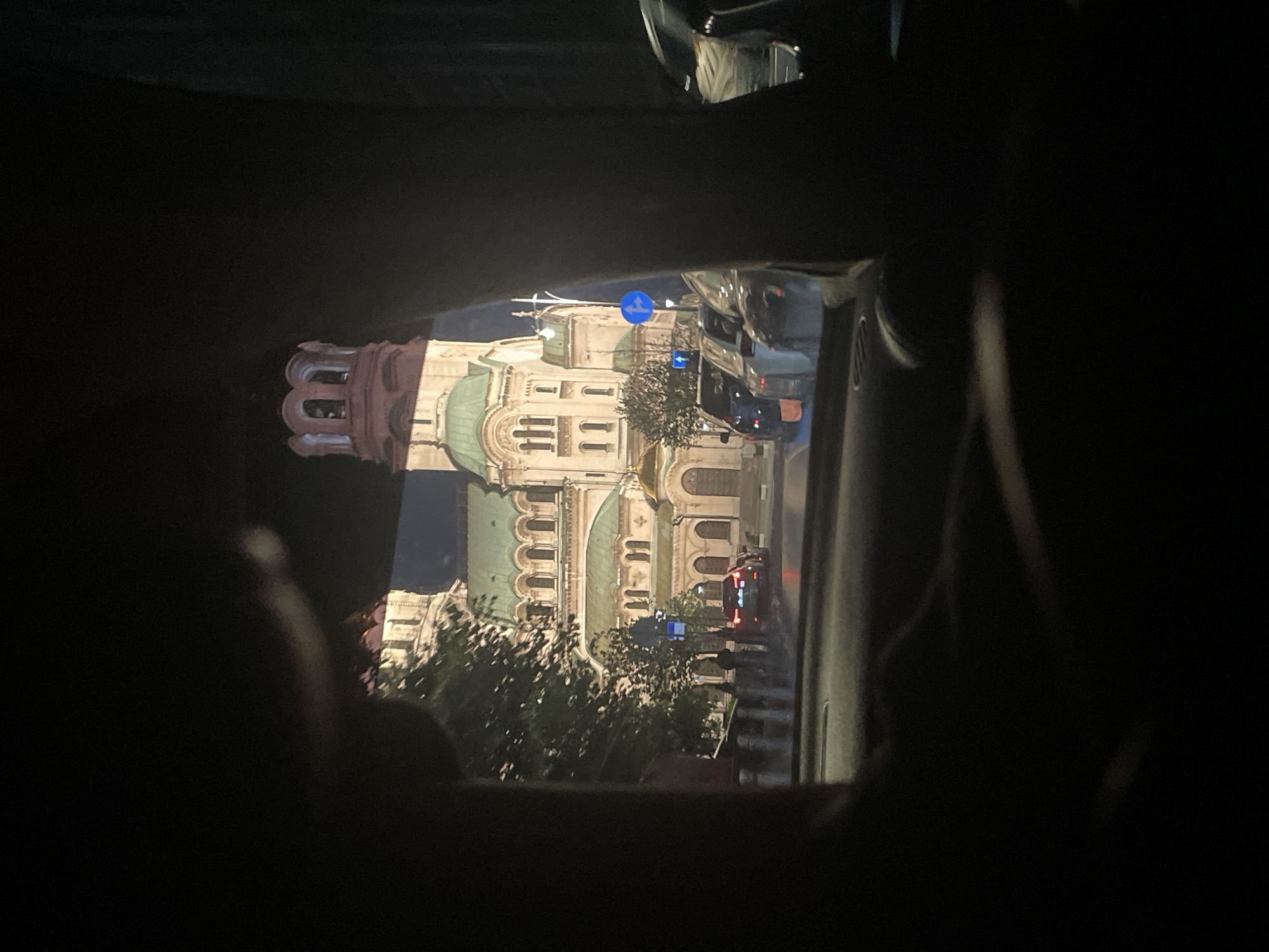 The image, taken from within a vehicle at night, shows a partial view of the St. Alexander Nevsky Cathedral in Sofia, Bulgaria. The cathedral is illuminated. Street signs and moving vehicles are visible. The view is framed by the interior of the vehicle, with dark portions obscuring much of the image. Trees are visible in the foreground in front of the cathedral.