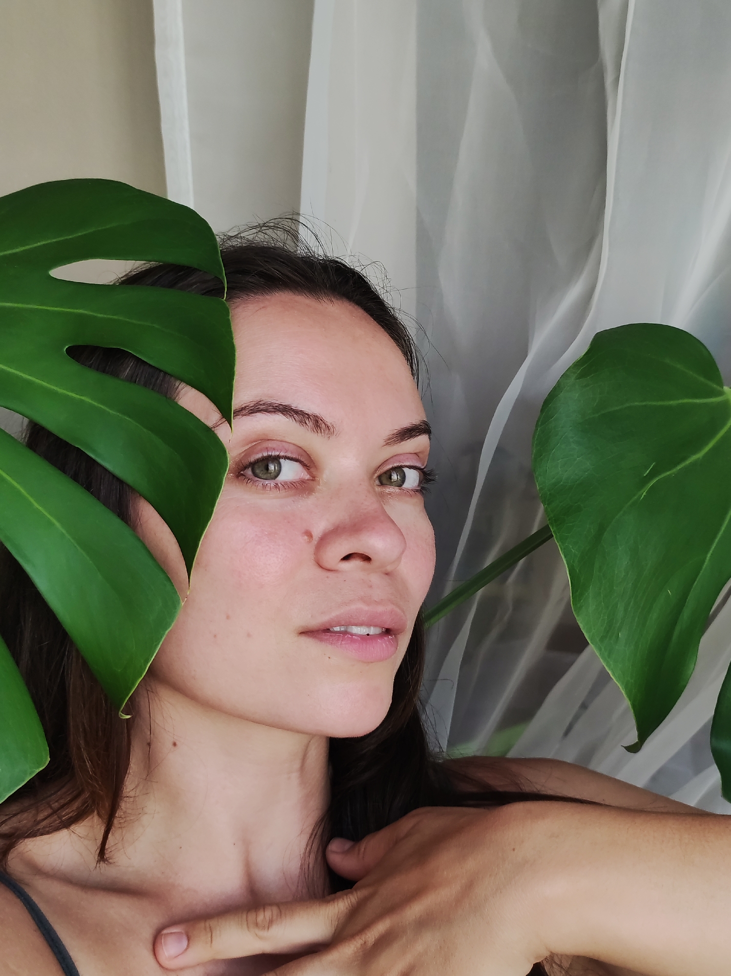 A close-up, medium shot shows a young woman's face peeking from behind large, vibrant green Monstera leaves. She has dark hair and fair skin with a light dusting of freckles across her nose and cheeks. Her eyes are a striking green-hazel, and she gazes slightly off-camera with a subtle, soft smile. Her left cheek is partially obscured by a large Monstera leaf, and another leaf frames the right side of her face. The background consists of a sheer, white, translucent curtain, creating a soft, diffused light. Her right hand is visible, resting on her collarbone, with her fingers gently extended. The image is set indoors, likely in a home environment in Chaikovsky, Russia, given the presence of houseplants and the domestic backdrop.