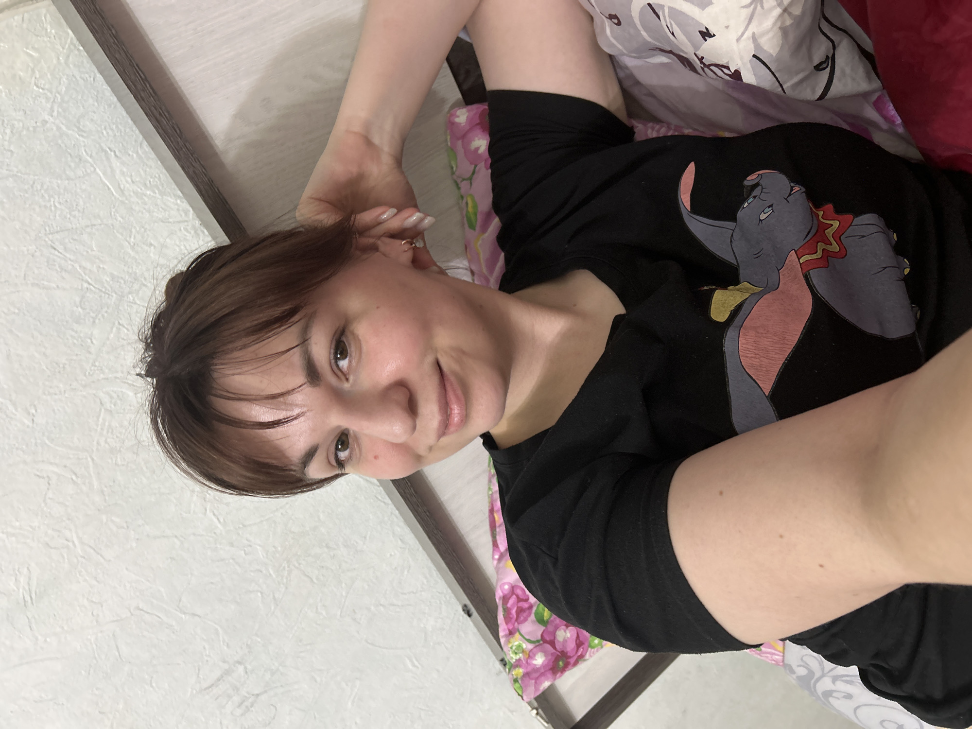 The image displays an adult female in an indoor setting, consistent with a bedroom. The specific location in the city of Фоки, Russia, cannot be independently verified from the visual information provided. The subject, an individual with brown hair and light skin, is positioned from the chest up, reclining or lying down, looking directly at the camera with a slight smile. She is wearing a black short-sleeved t-shirt featuring a graphic illustration of the cartoon character Dumbo. A small hoop earring is visible in her right ear, and her fingernails display a light-colored polish. Her left arm is bent, with her hand positioned near her head. The background consists of a light-colored, textured wall. Elements of a dark-colored bed frame or headboard are partially visible behind her head. Bedding is present: a pink fabric with a floral pattern is visible on the lower left, and a grey and white patterned fabric is visible on the lower right, with an additional reddish fabric also present on the far right.