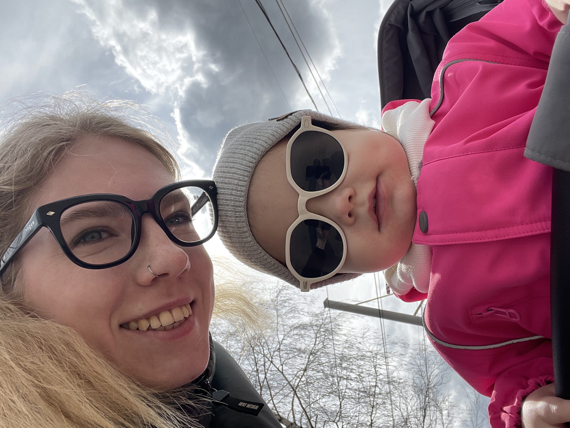 A low-angle outdoor selfie captures a woman and a young child against a cloudy sky with patches of bright sunlight, set in Izhevsk, Russia. The woman, positioned on the left, has blonde hair, wears black-rimmed glasses with "Vogue" visible on the arm, and a silver nose ring. She is smiling broadly. Her dark jacket features a fur-lined collar, and a tag on the front reads "BEST DESIGN."
To her right, partially obscured and tilted, is a young child or baby. The child wears a light grey knit beanie and stylish round, light-framed sunglasses, and smiles slightly. They are dressed in a bright pink jacket with reflective piping and a zippered pocket, layered over a white garment. A grey stroller or carrier is partially visible behind the child.
The background shows bare tree branches, suggesting a late winter or early spring season, and overhead power lines. The sky is largely overcast but allows some sunlight through. Both subjects appear to be in good spirits, conveying a happy and warm moment despite the cool weather indicated by their attire.