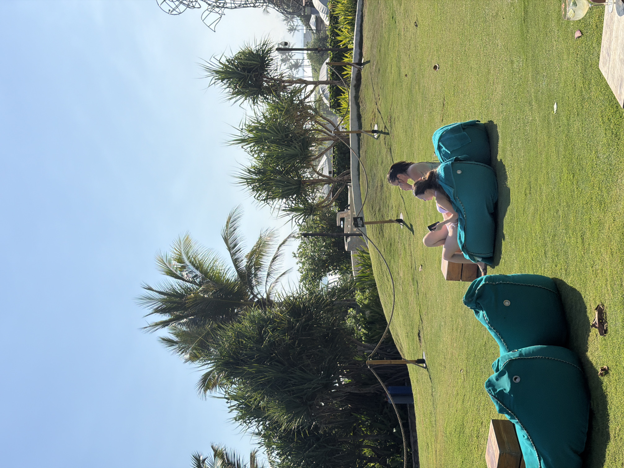 The image depicts an outdoor seating area with lush green grass under a clear blue sky. In the foreground, two large teal-colored beanbag chairs are placed on the lawn. To the right of these chairs, two women are relaxing. The woman closer to the camera is wearing a light-colored swimsuit and looking at her phone, while the woman behind her is wearing a darker top and looking upwards. Behind the women, another teal beanbag chair is visible. In the background, several palm trees and other tropical vegetation are present, suggesting a warm, possibly coastal, environment. A rope barrier with wooden posts is strung across a portion of the lawn. Further in the background, structures and umbrellas suggest a resort or a hotel. The image does not contain any discernible elements that would allow for its specific location within the city of Kediri, Indonesia.