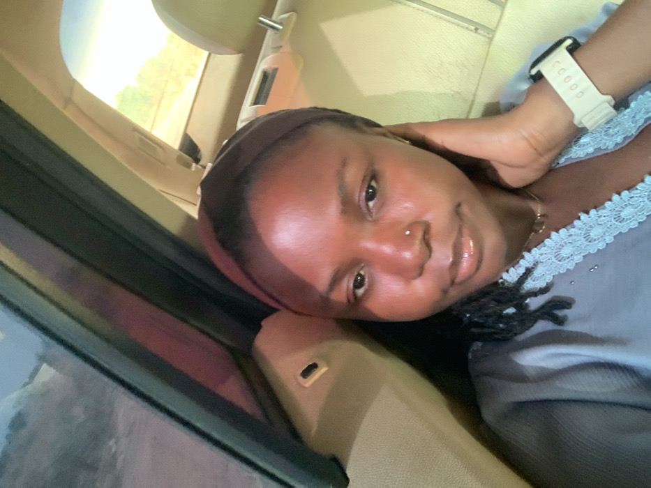 The image depicts a young woman seated in the passenger compartment of a vehicle, appearing to be the back seat, located in Gadam, Nigeria. She has dark skin, dark braided hair visible on her left shoulder, and is wearing a dark brown or black headwrap. A small metallic stud is visible in her left nostril. Her expression is neutral, and her lips appear moisturized. She is wearing a light blue garment with a V-neckline adorned with light blue lace trim. A delicate necklace with small dark charms is visible around her neck. On her left wrist, she wears a light beige or off-white digital smartwatch. Her left hand is raised, with fingers touching the side of her face or hair near her left ear. The vehicle's interior features light-colored (beige or tan) upholstery and plastic trim on the door panel and seat headrest. Through the windows, blurred natural light and faint outlines of an outdoor environment, possibly a dirt road and distant foliage, are discernible.