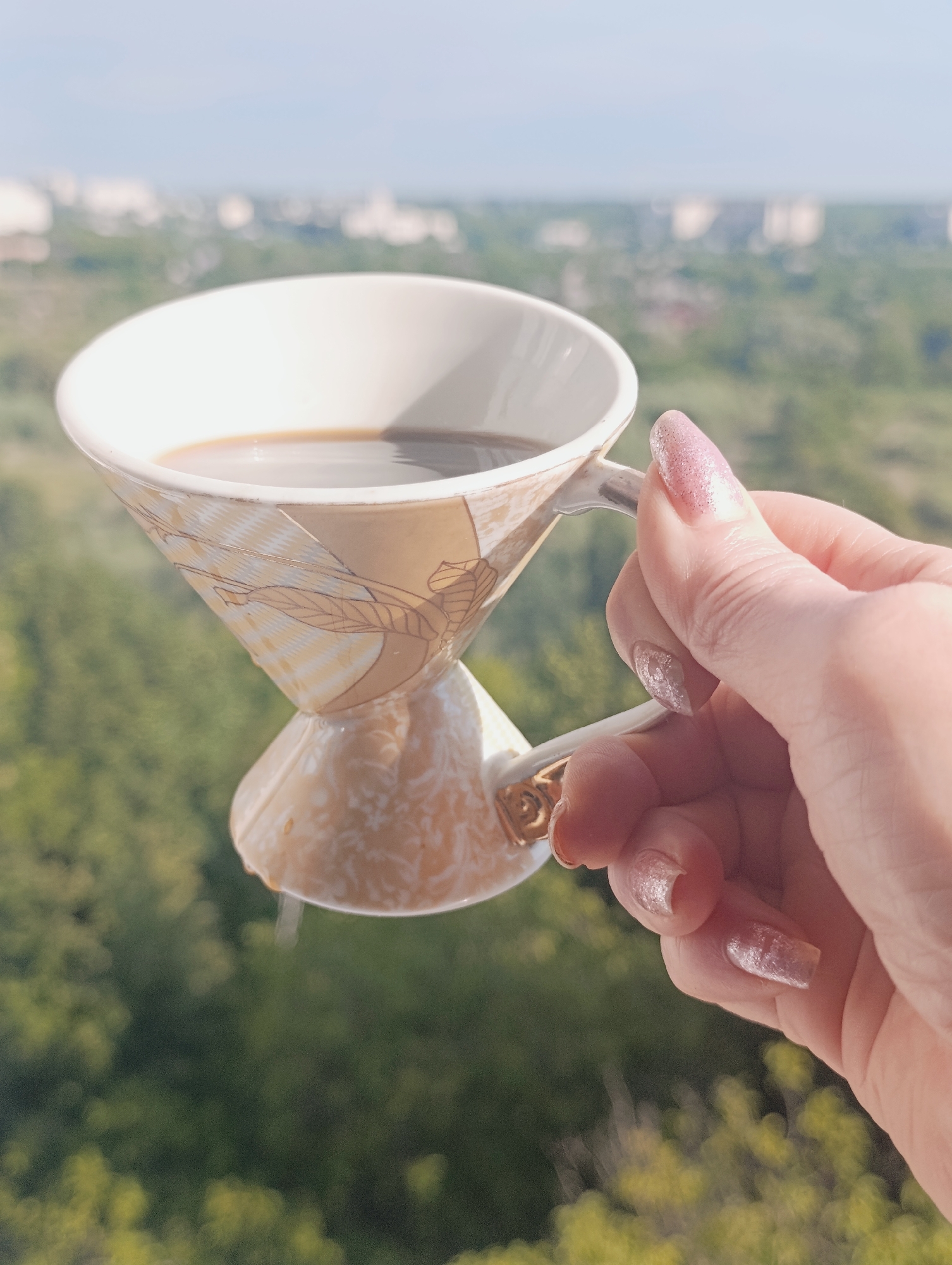 In an outdoor setting with a blurred cityscape and green foliage in the background, a person's hand with glittery fingernails holds an ornate, uniquely shaped coffee cup filled with dark liquid. The cup is a pale beige with a pattern of stylized leaves and stems, and it has a narrow base and a wider top. The hand is positioned to the right of the cup, with the fingers gripping the handle. The environment suggests a clear day with buildings and trees visible in the distance, possibly indicating a residential or urban area. The focal point of the image is the cup and the hand holding it. The location is identified as Bila Tserkva, Ukraine.
