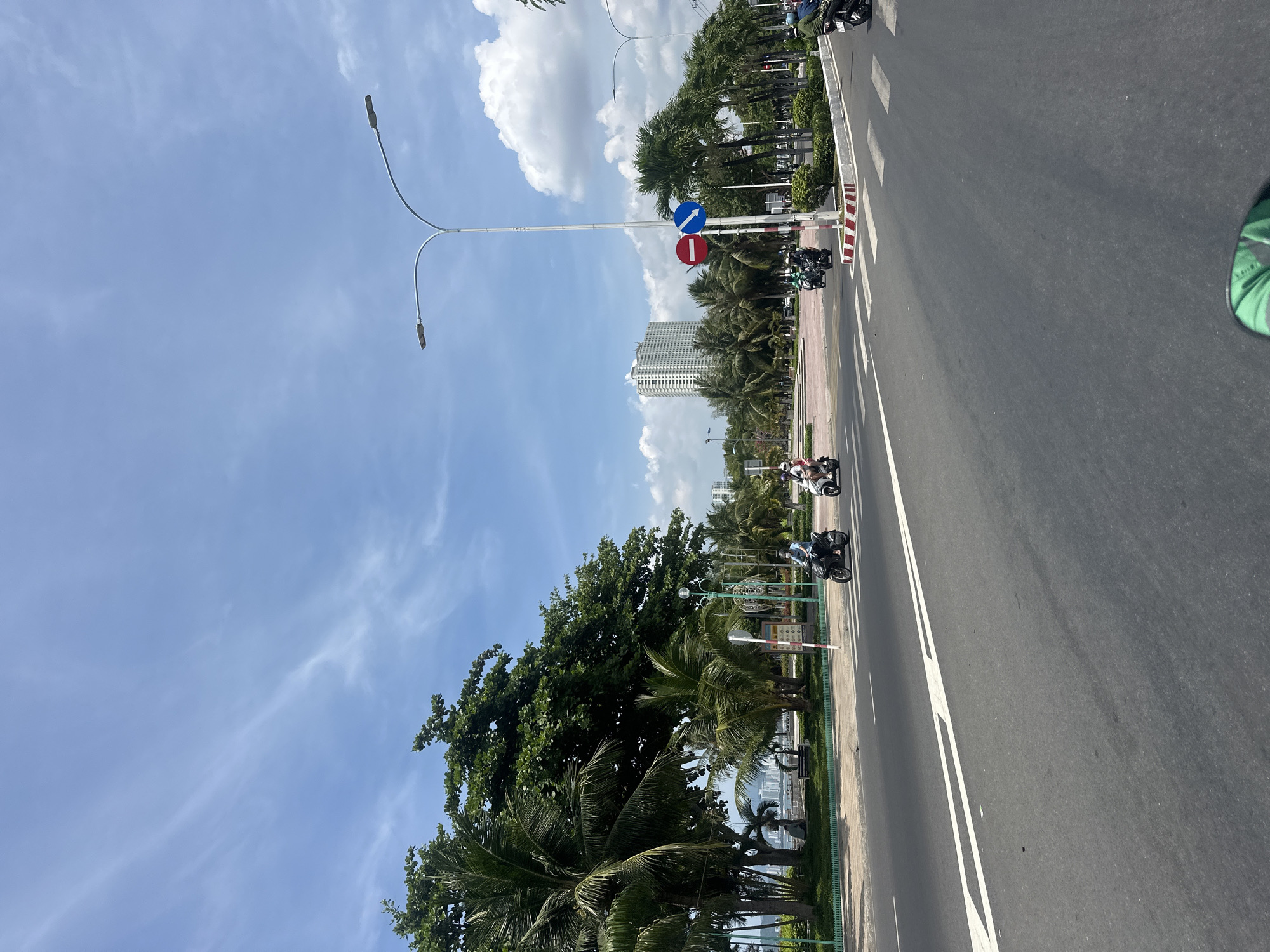 The image depicts a sunny day in Bac Nha Trang Ward, Vietnam, with a street scene focused on a road and a pedestrian crossing. A white road marking with an arrow indicates the direction of travel. Several motorbikes are visible, with riders in motion, suggesting traffic. On the side of the road, palm trees and other lush greenery line the area, creating a tropical atmosphere. In the background, a tall white building stands out against the blue sky, interspersed with white clouds. A pedestrian crossing with striped markings is present, and traffic signs, including a red and white circular sign with a horizontal bar and a blue downward-pointing arrow, are also visible. The overall scene suggests an urban coastal environment with active transportation and clear weather.