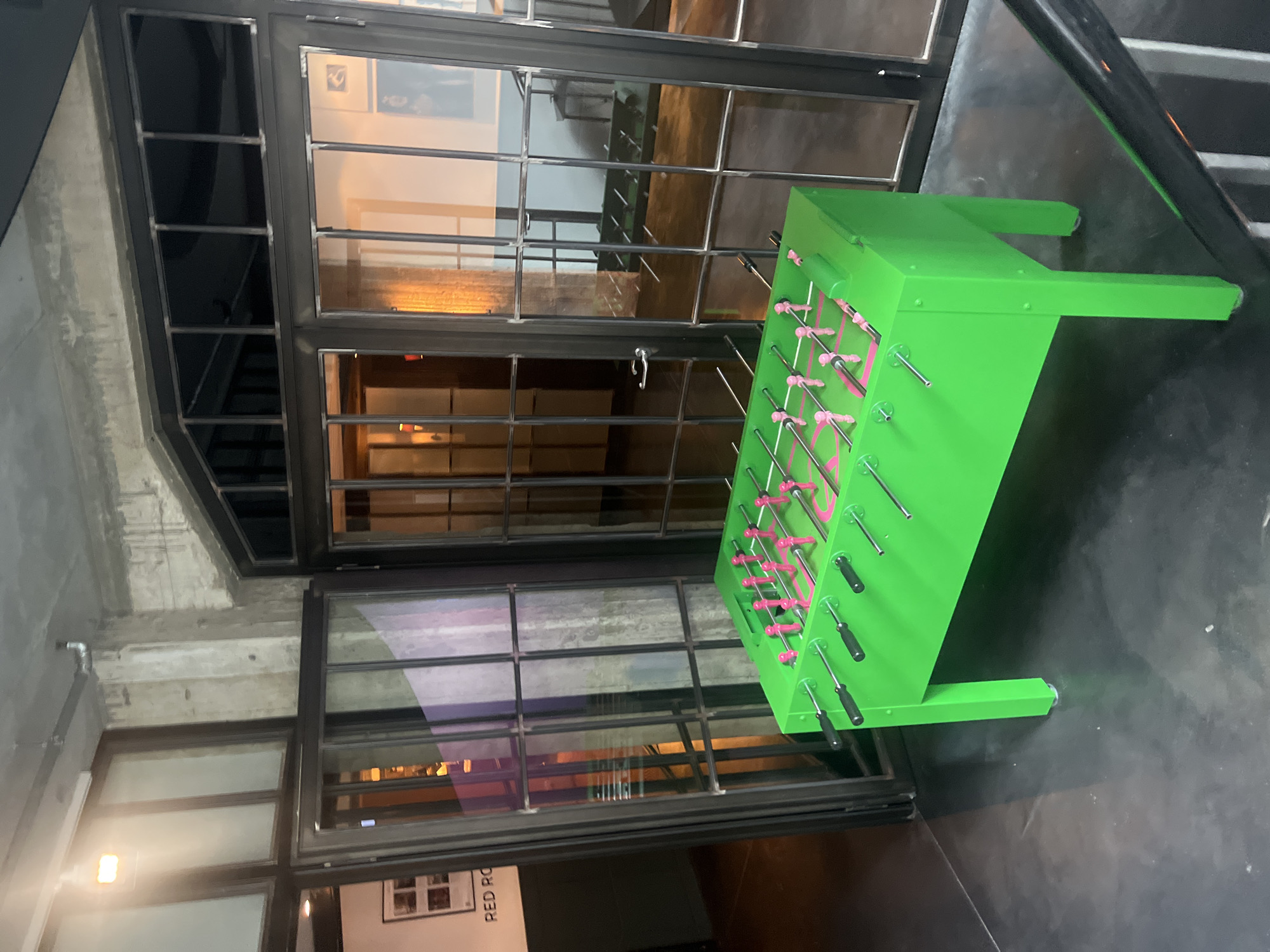A bright green foosball table with pink player figures is centrally positioned on a dark, polished concrete floor. The table is in the foreground, occupying the lower half of the image. Behind the foosball table are large, industrial-style glass doors with dark metal frames. Through the glass doors, a dimly lit interior space is visible, featuring concrete pillars and a few scattered objects. To the left of the foosball table, a partially open glass door reveals a sign on the wall that reads "RED RO". The ceiling appears to be exposed concrete with visible pipes and industrial lighting. The overall environment suggests an indoor venue, possibly a bar or entertainment space, located in Berlin, Germany.