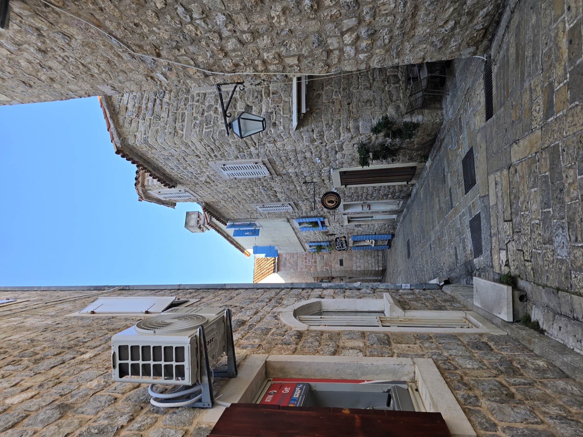 The image depicts a narrow cobblestone street in the Old Town of Budva, Montenegro. The street is flanked by old stone buildings with red-tiled roofs. On the left side, a building has an ATM machine visible. Further down the street, another building features blue shutters. On the right side, a stone building has a traditional street lamp mounted on its exterior. In the distance, another building with blue shutters is visible. The street is empty, with no people present. The sky is clear and blue.