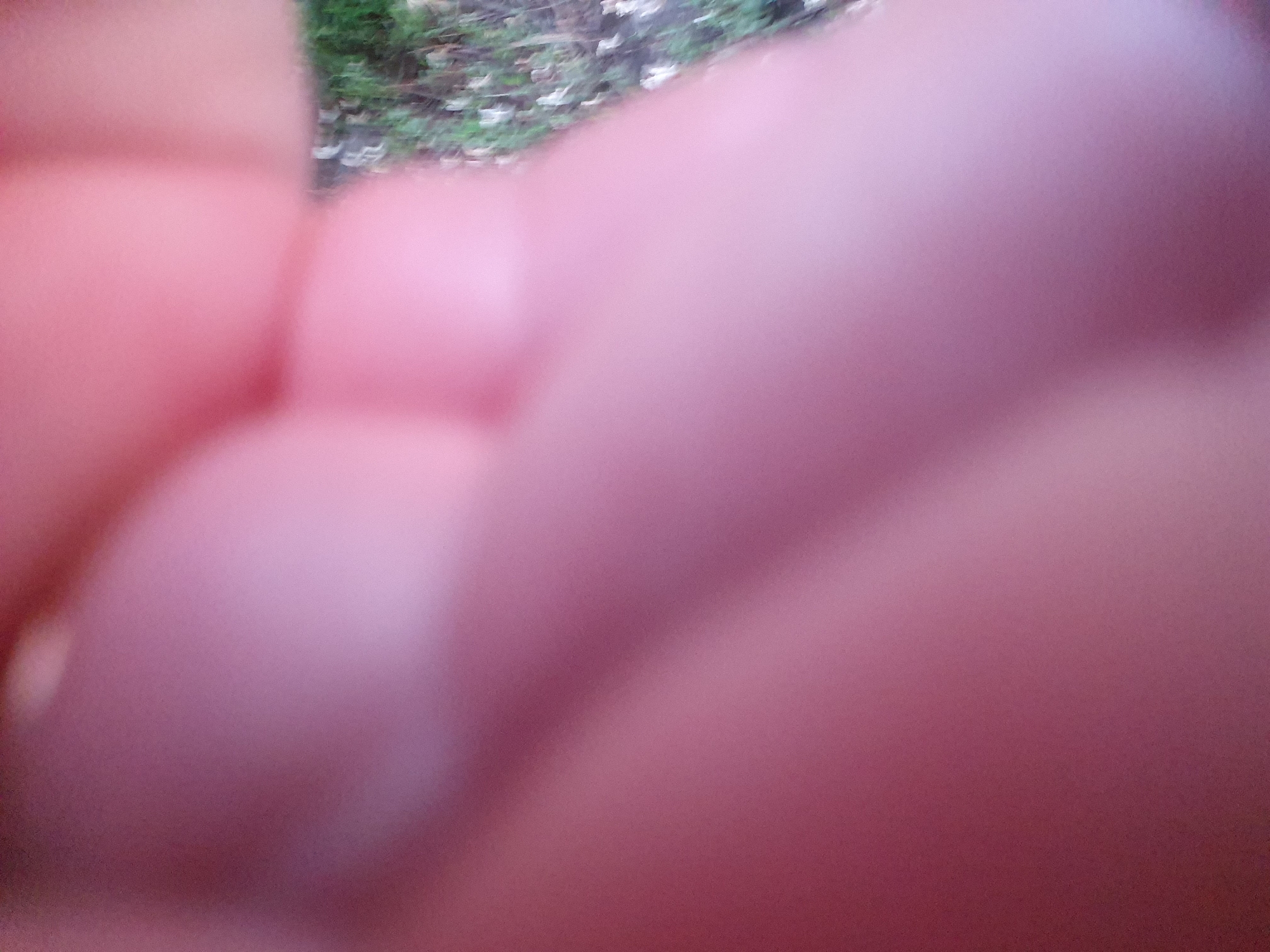 The image is a close-up, out-of-focus shot of what appears to be a person's hand and arm, with the background showing some green foliage and possibly a dirt or gravel path. The dominant colors are soft pinks and purples from the skin, transitioning to muted greens and browns in the background. The lighting suggests it might be outdoors during the day, but the blurriness prevents any specific identification of the location or any precise details of the hand or arm.