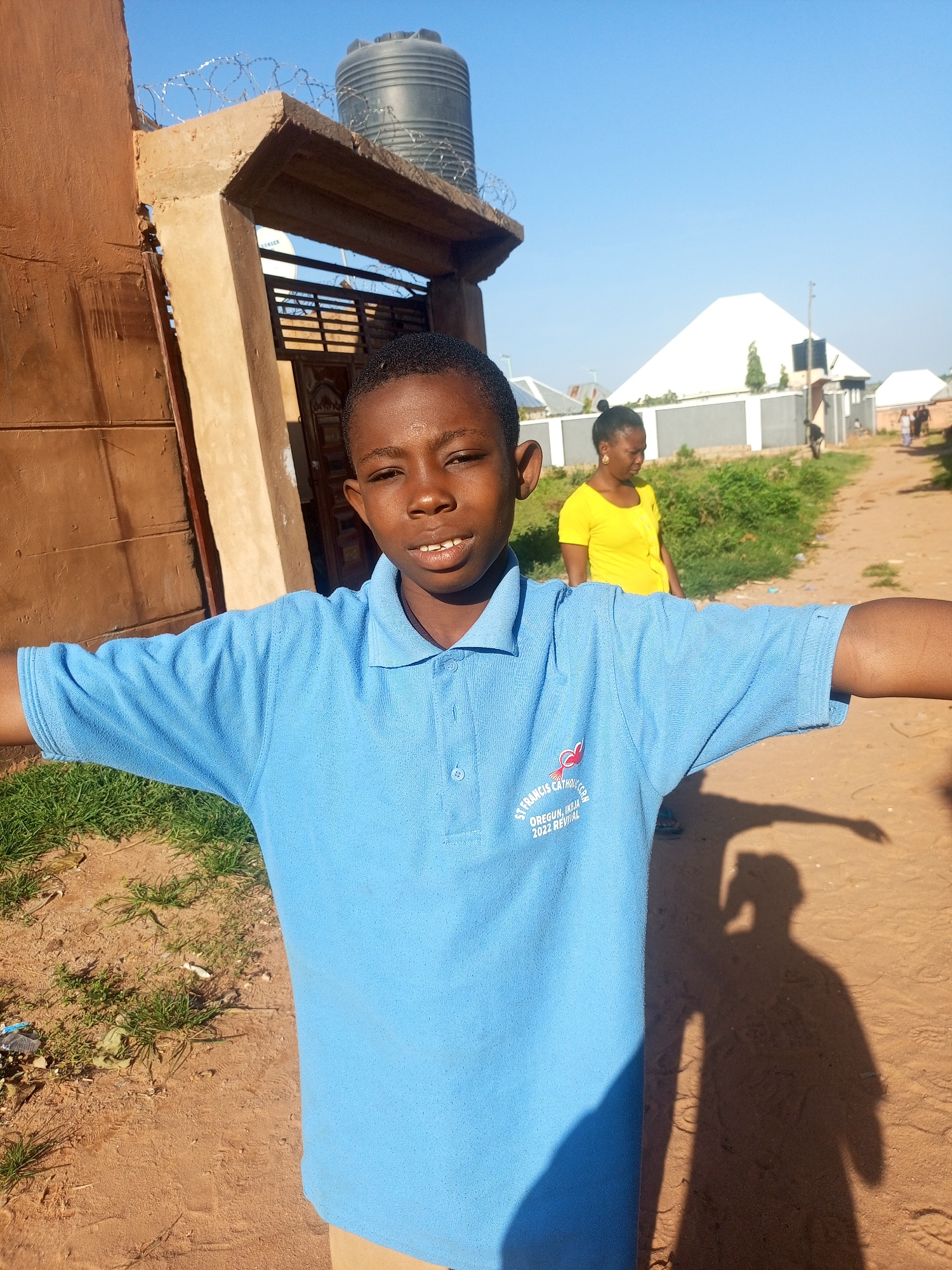 In Tempure, Nigeria, a young boy in a light blue polo shirt with a school emblem stands with his arms outstretched. He is positioned in the foreground, facing the camera with a slight smile and open mouth. Behind him, a woman in a yellow t-shirt walks away from the camera, and other figures are visible in the distance. The environment appears to be a dusty, unpaved road, with a building constructed of brown material and a concrete archway to the left. A large black water tank is mounted on top of the building, surrounded by barbed wire. In the background, several houses with white roofs and fences are visible, along with scattered greenery. The sky is clear and blue. The sun casts a prominent shadow of the boy on the ground.