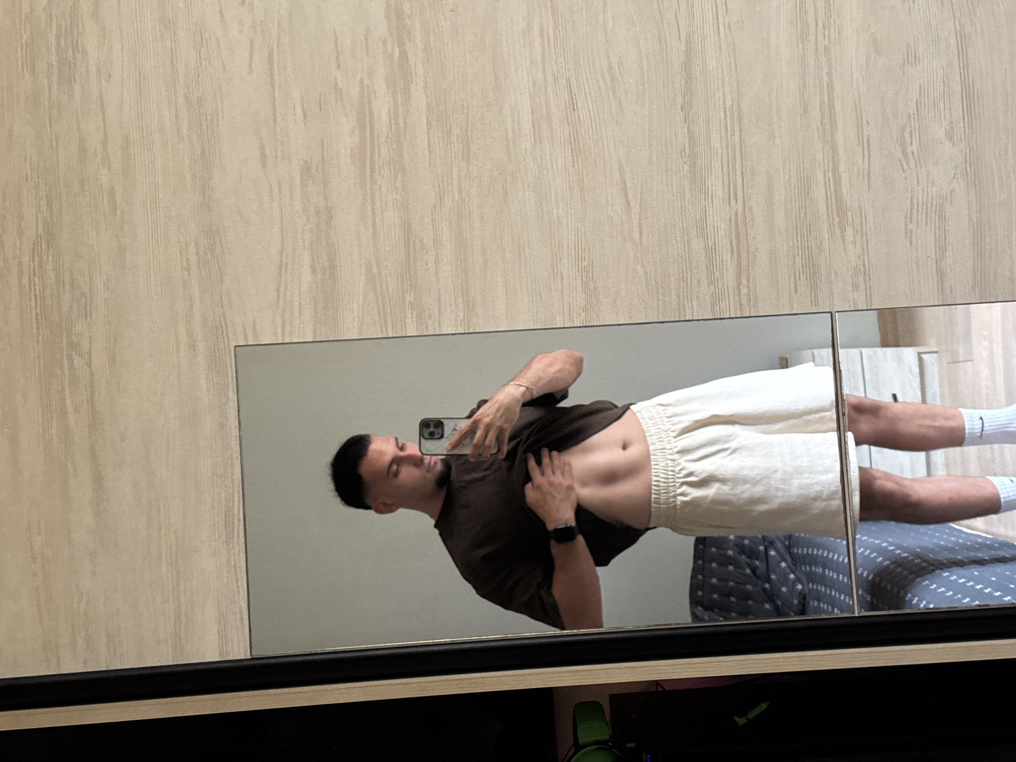 A person is standing in front of a mirror, taking a selfie. They are wearing a dark t-shirt and light-colored shorts, with white socks. The person has short dark hair and is looking down at their phone, which they are holding in their right hand. Their left hand is on their stomach, showing their abs. The background is a light-colored wall with a wood-grain pattern. The mirror reflects the person and the room behind them, which appears to be a bedroom with a bed and a patterned comforter. The image is taken from a slightly high angle. There is no visible text in the image. The lighting suggests it is daytime.